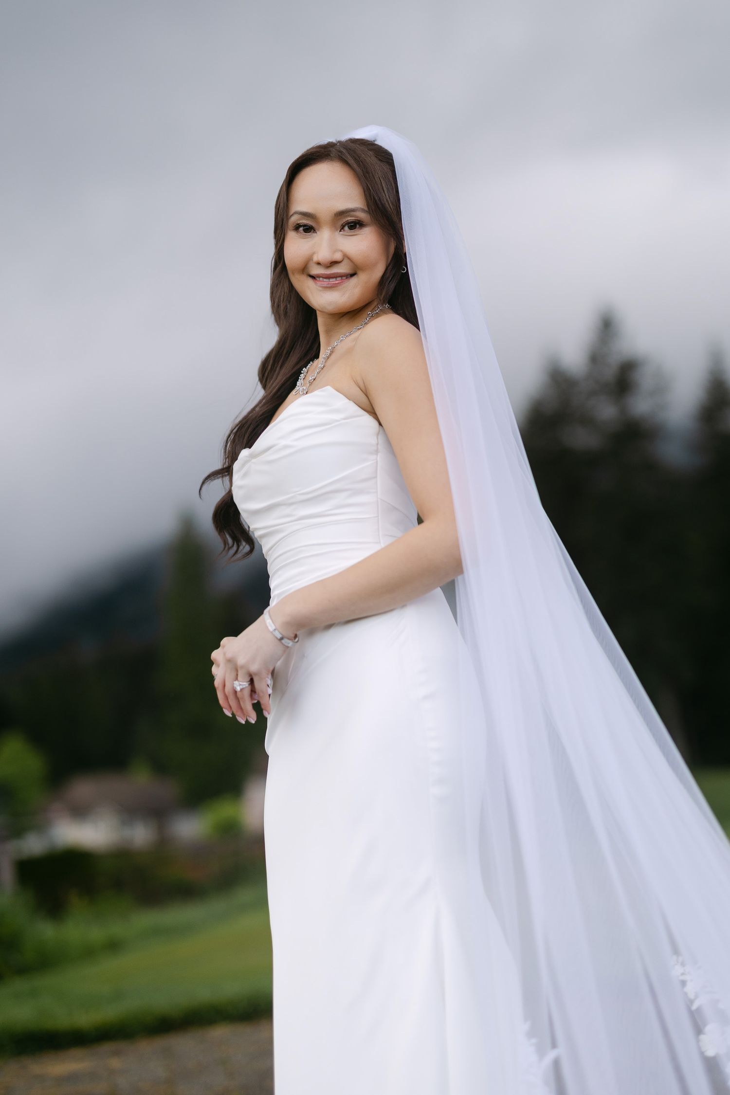 Smiling bride in white strapless gown and veil poses outdoors with evergreen trees and cloudy sky.