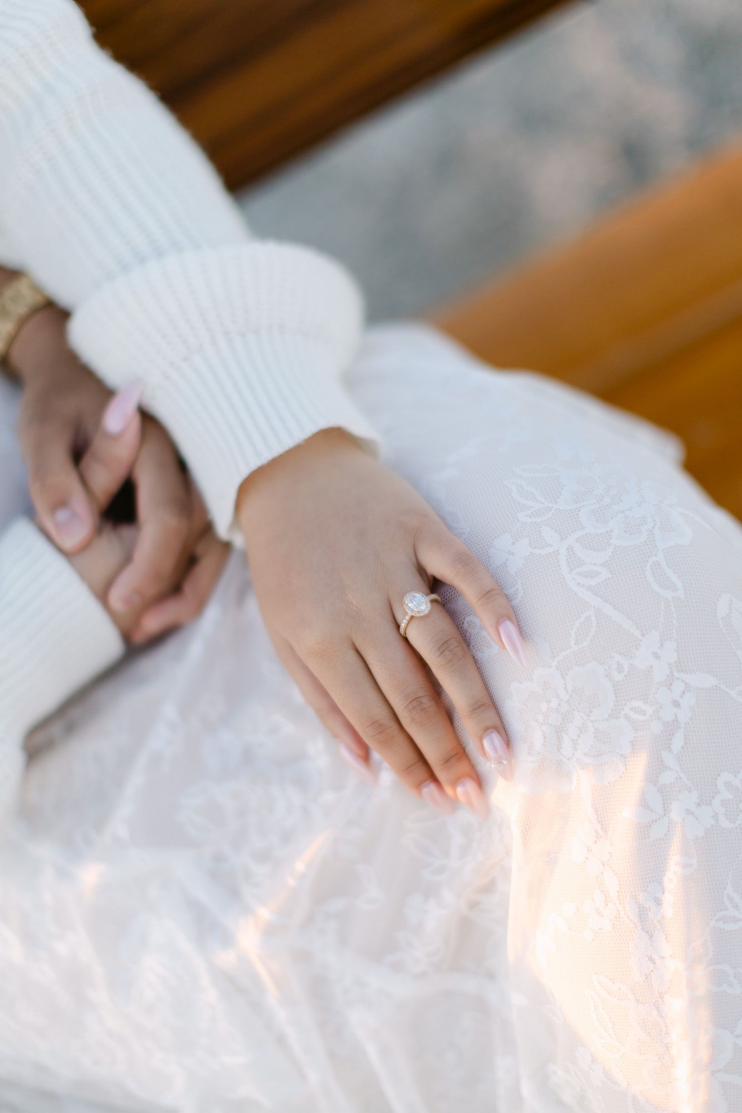 Close-up of a woman's hand wearing a diamond engagement ring resting on her white lace dress.