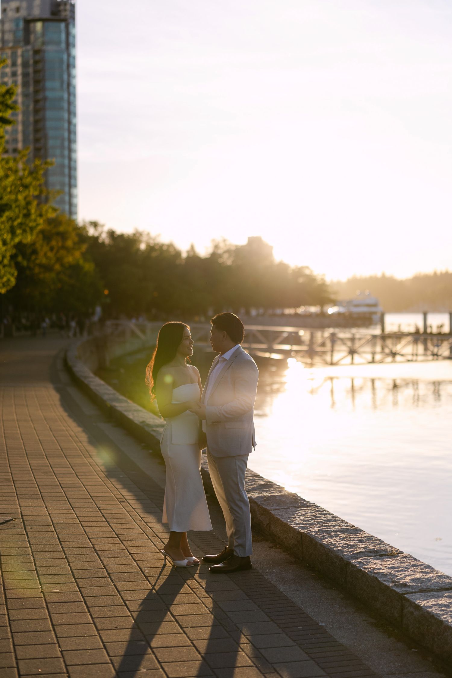 Couple embracing on a brick waterfront promenade at golden hour sunset, with a marina, trees and city high-rises in the background.