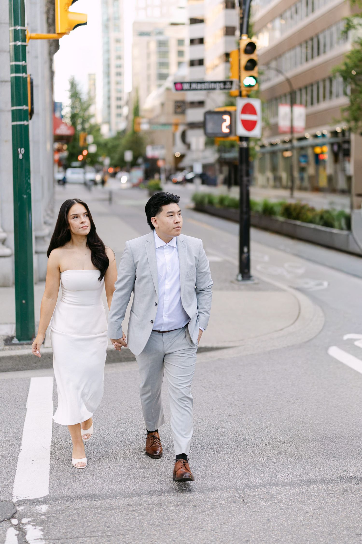 Engaged couple holding hands while crossing a downtown city street, she in a white strapless dress and he in a light gray suit.