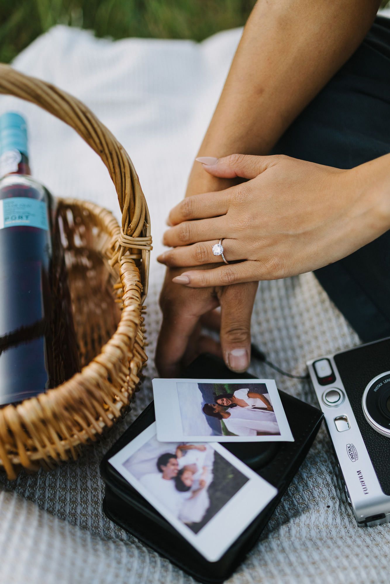 Close-up of a hand with a diamond ring resting on instant photos beside an instant camera and a wicker basket with a wine bottle.