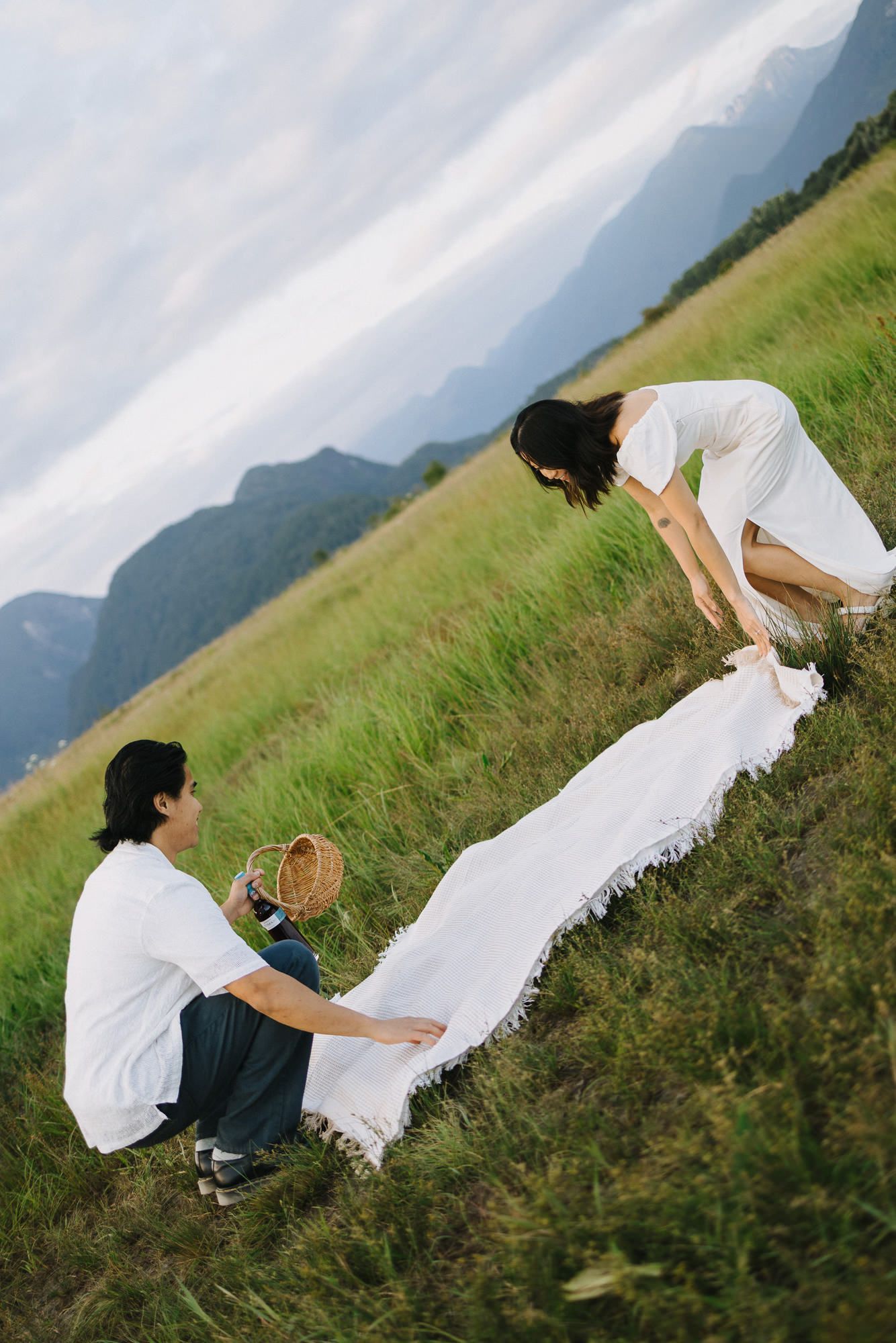 Couple setting up a picnic blanket on a grassy hillside with mountain views, woman in a white dress leaning over.
