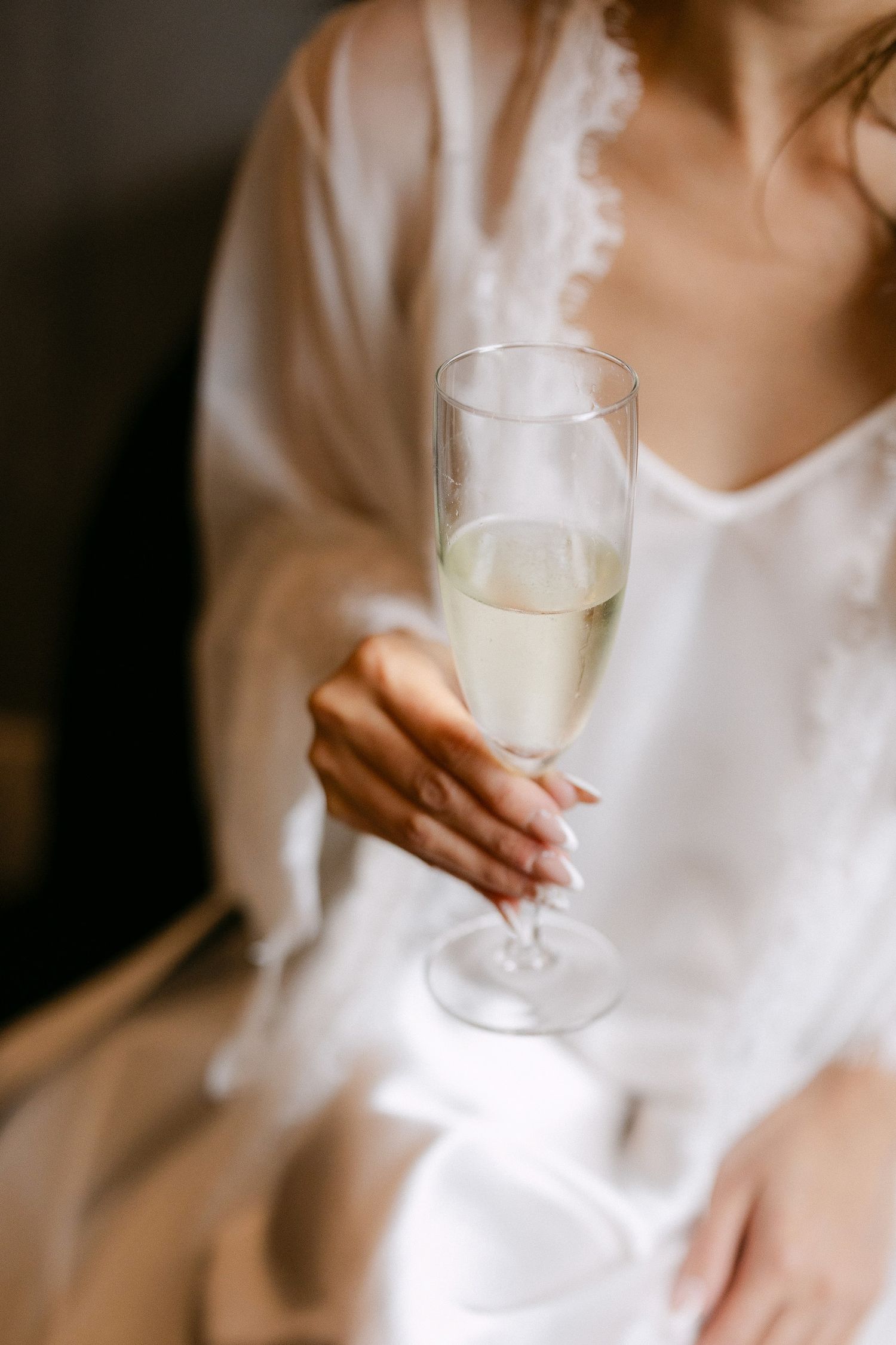 Bride in white silk robe holding a champagne flute during wedding morning preparations.