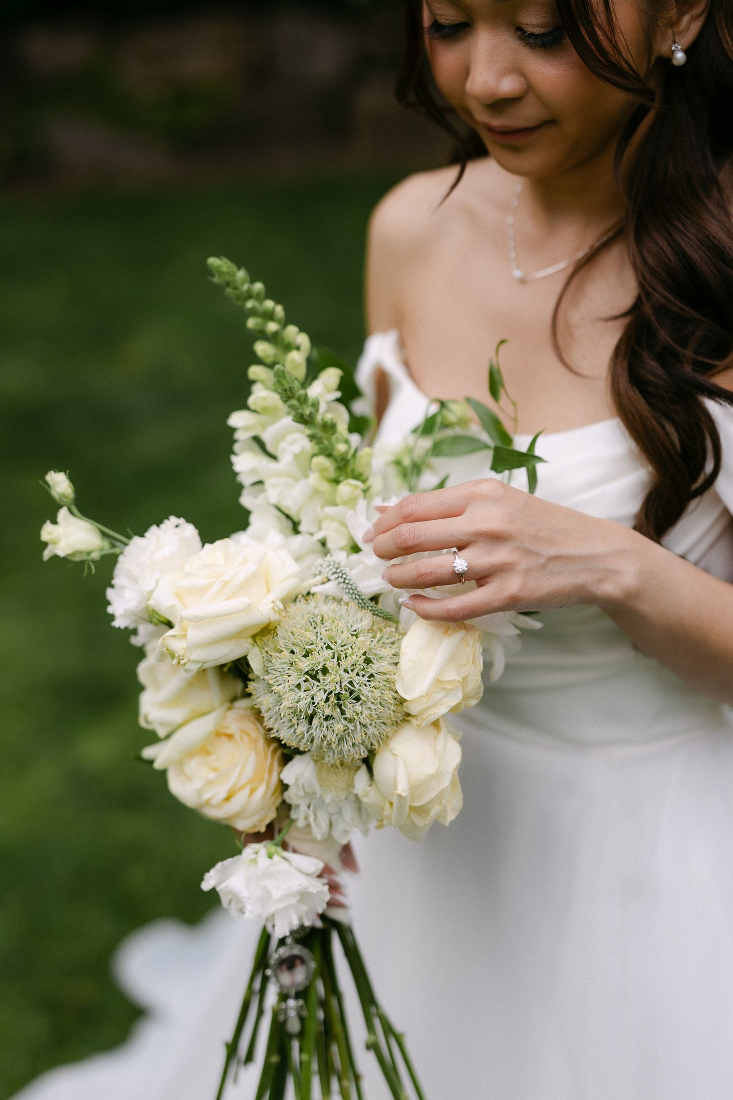 Bride in white gown holds elegant bouquet of ivory roses and green allium flowers, diamond ring visible.