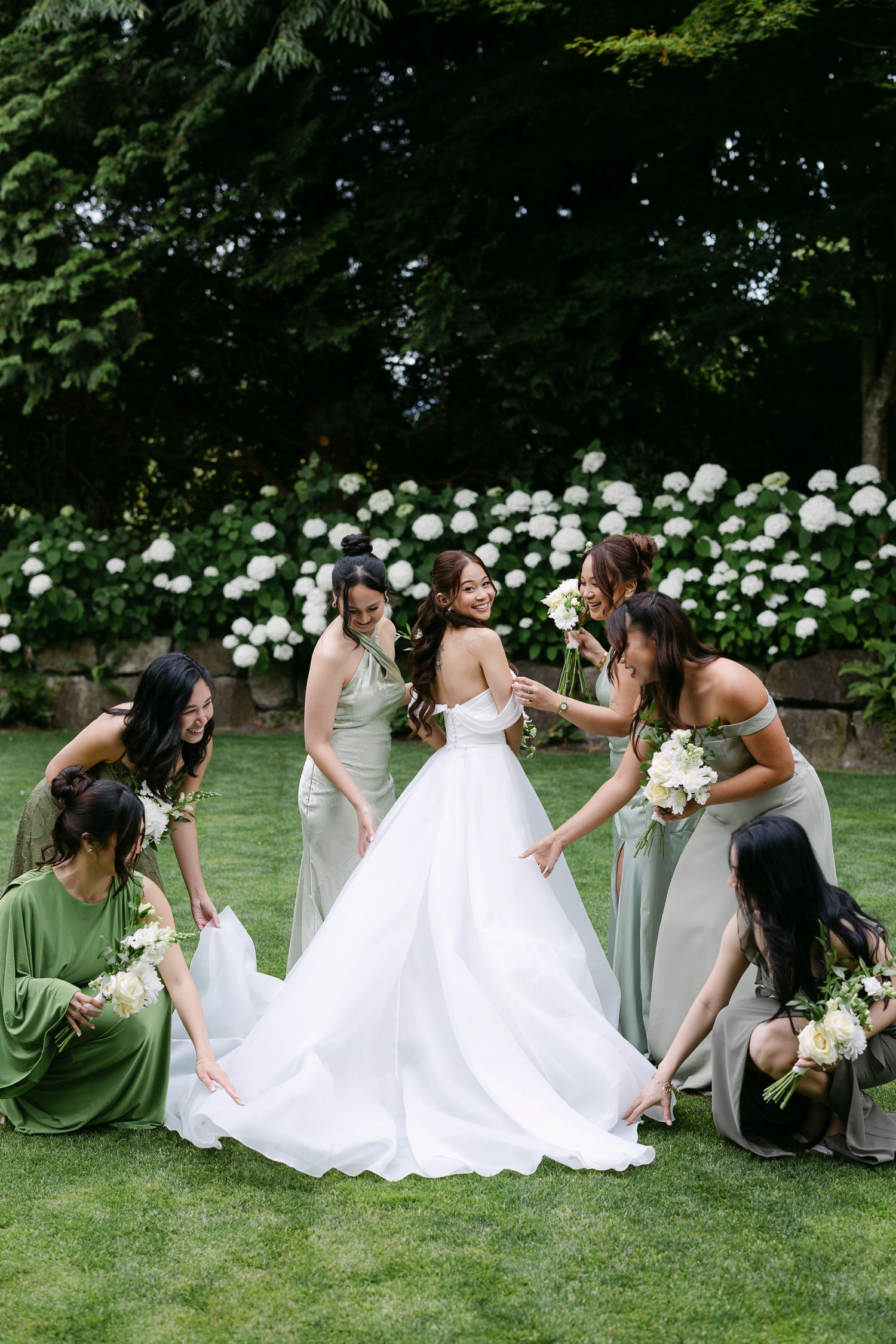 Bride in white ball gown surrounded by bridesmaids in sage green dresses arranging her train on a lawn.