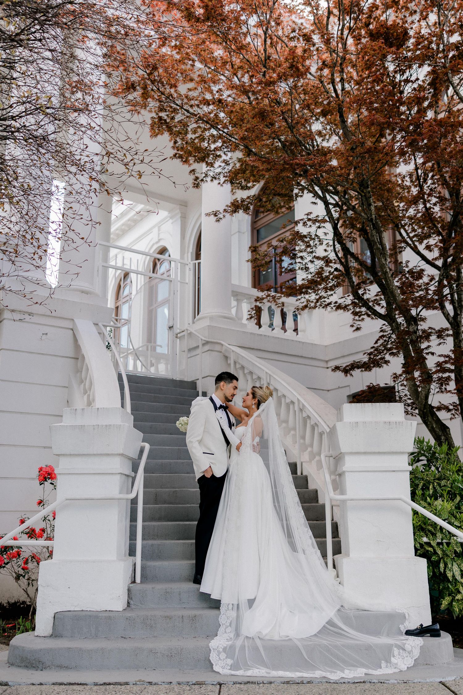 A couple shares a romantic moment on elegant white marble stairs with autumn foliage in the background.