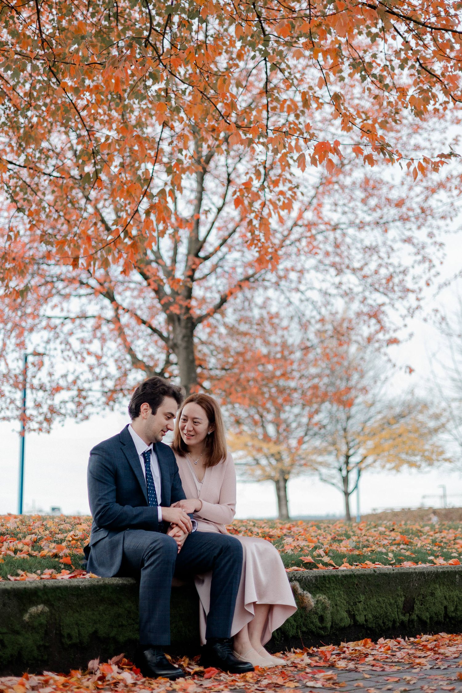 A couple sits together on a stone wall surrounded by autumn trees with vibrant orange leaves falling around them.