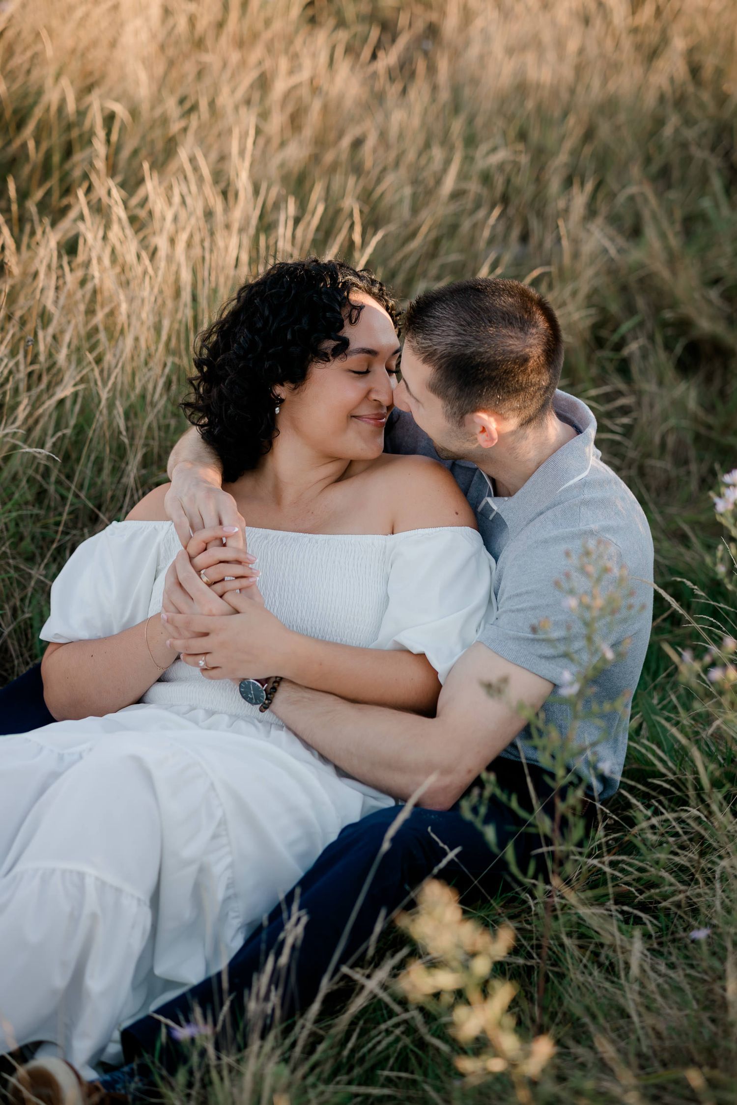 A romantic couple embraces in a field of tall grass during a sunset photoshoot.