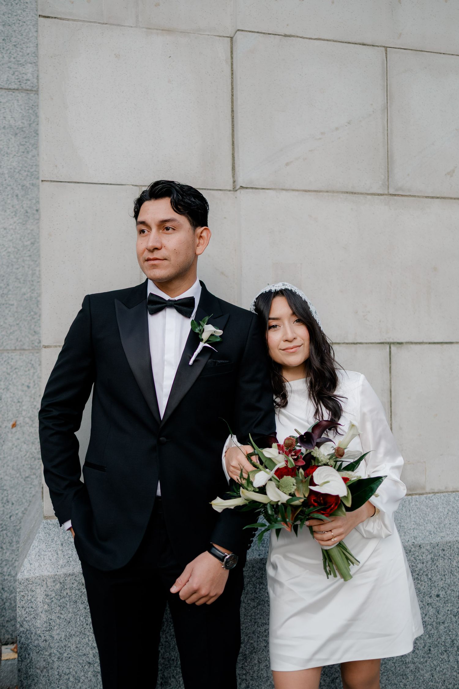 A couple in wedding attire poses against a light stone wall with a romantic floral bouquet.