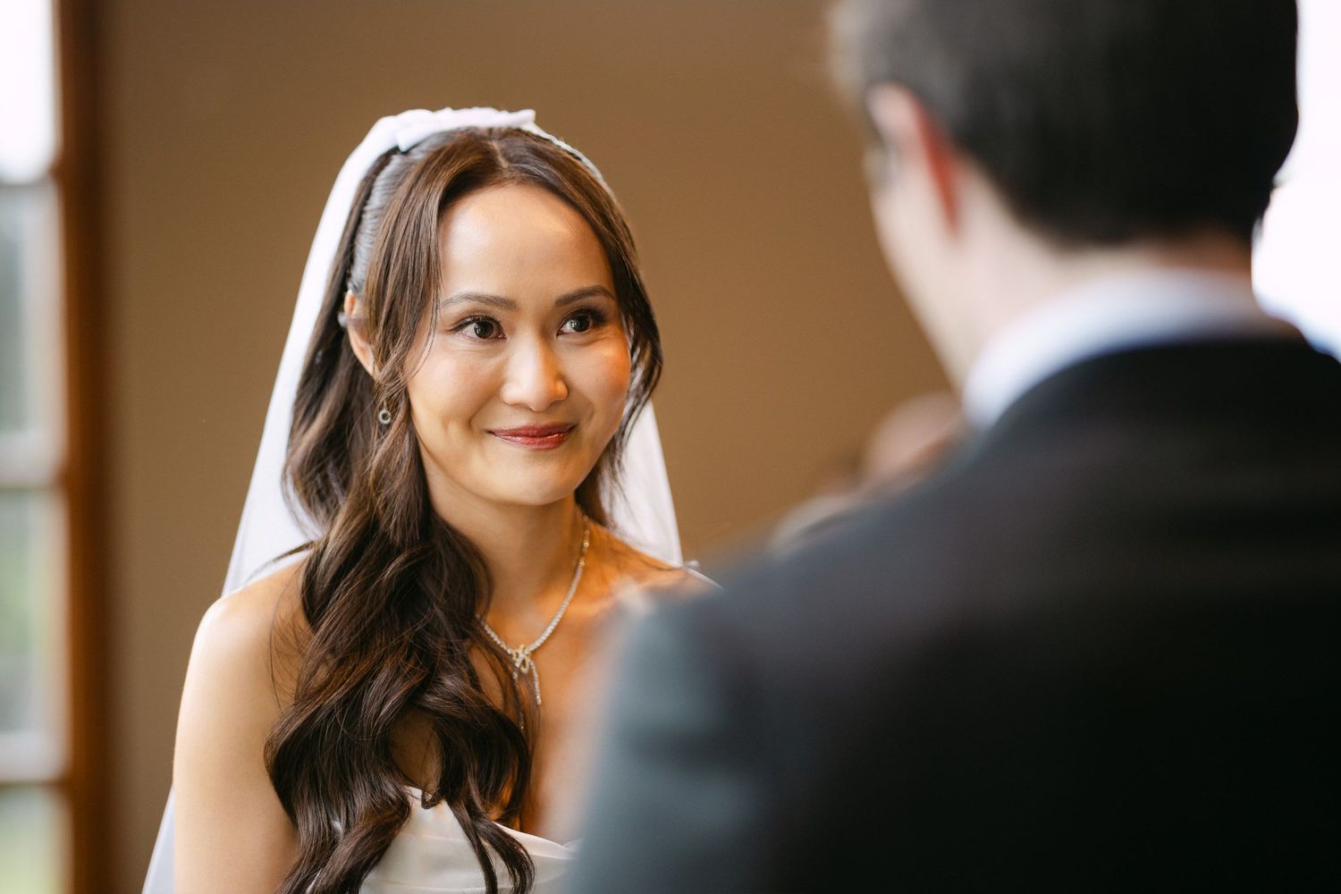Bride in white veil smiling warmly at groom during wedding ceremony indoors.