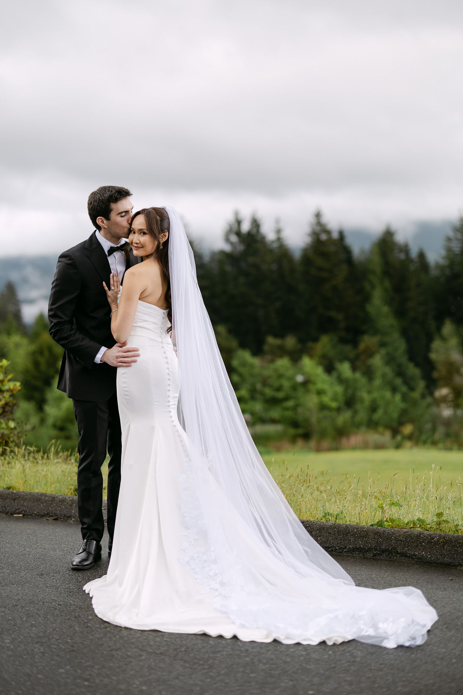 A bride in a white fitted gown with long veil embraces her groom in a black tuxedo outdoors with lush greenery.
