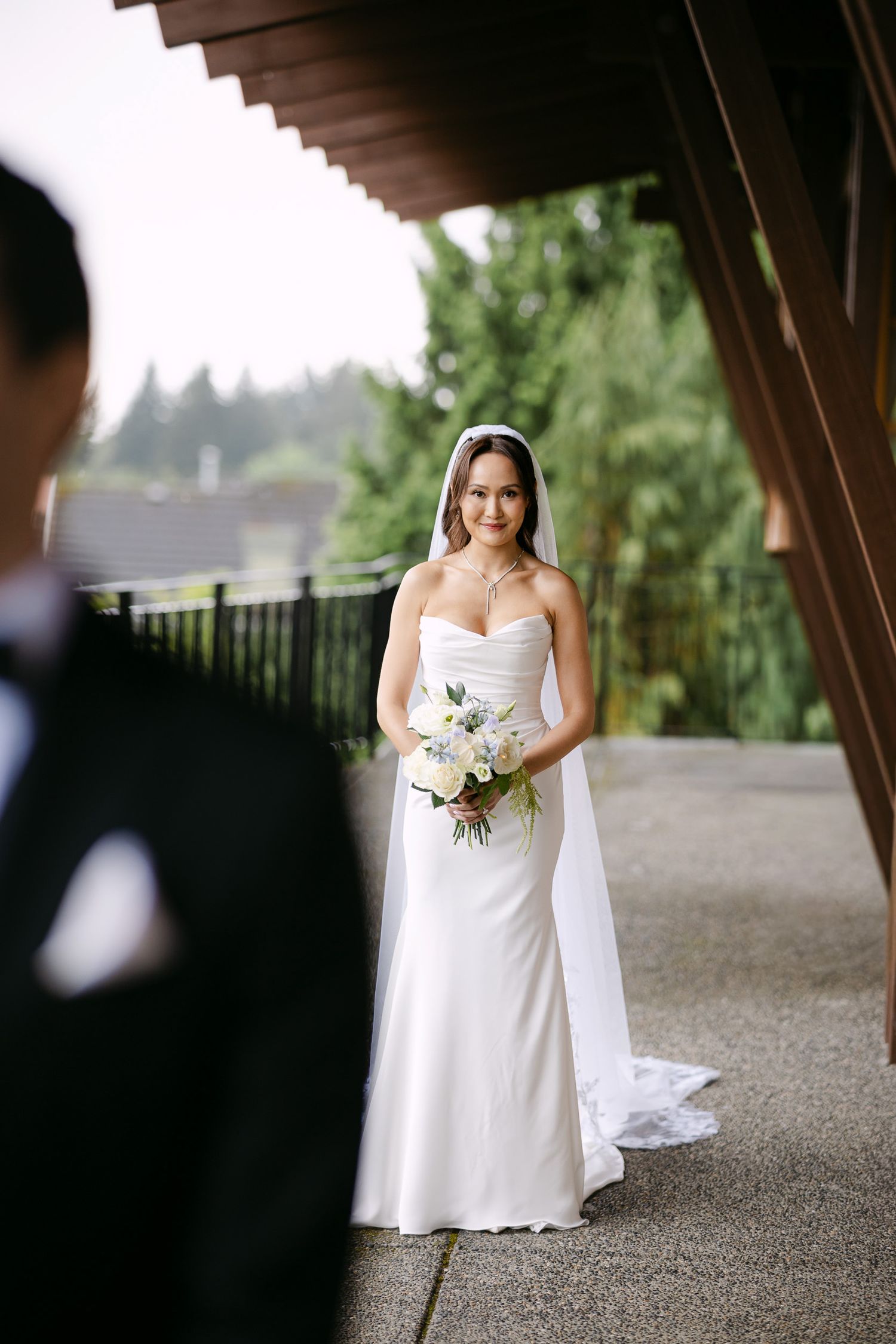 Bride in strapless white gown and veil holds bouquet outdoors, with groom in tuxedo in foreground.