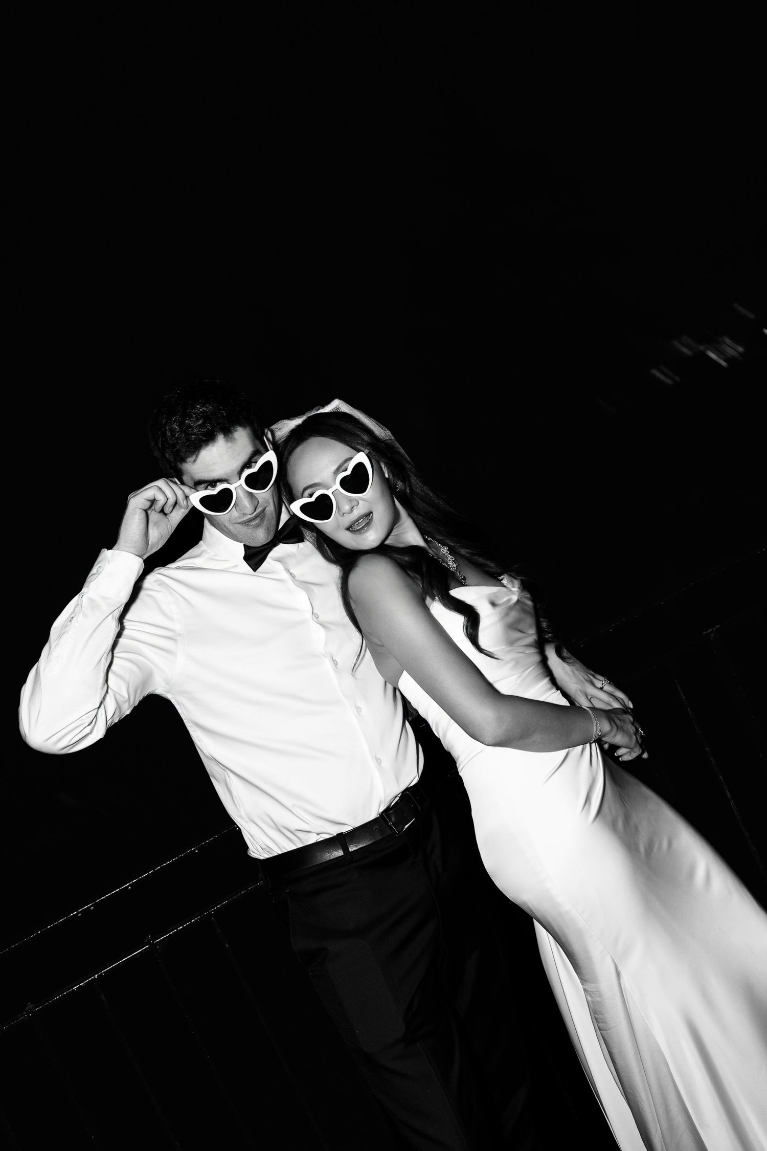 Black and white photo of an elegant couple in white outfits wearing heart-shaped sunglasses, posing together at night.