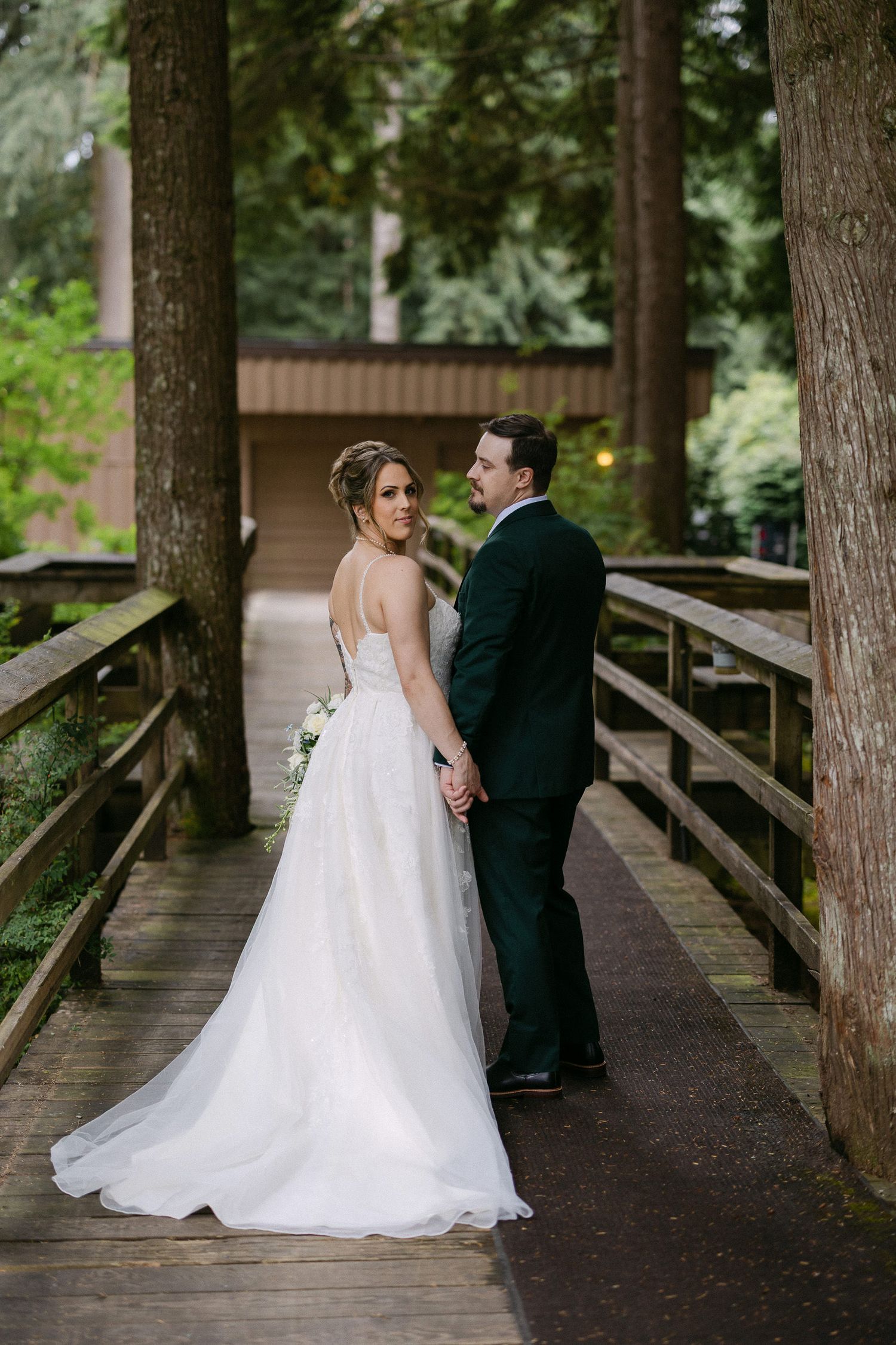 Bride and groom holding hands on a wooden bridge surrounded by tall trees in a lush forest wedding setting.