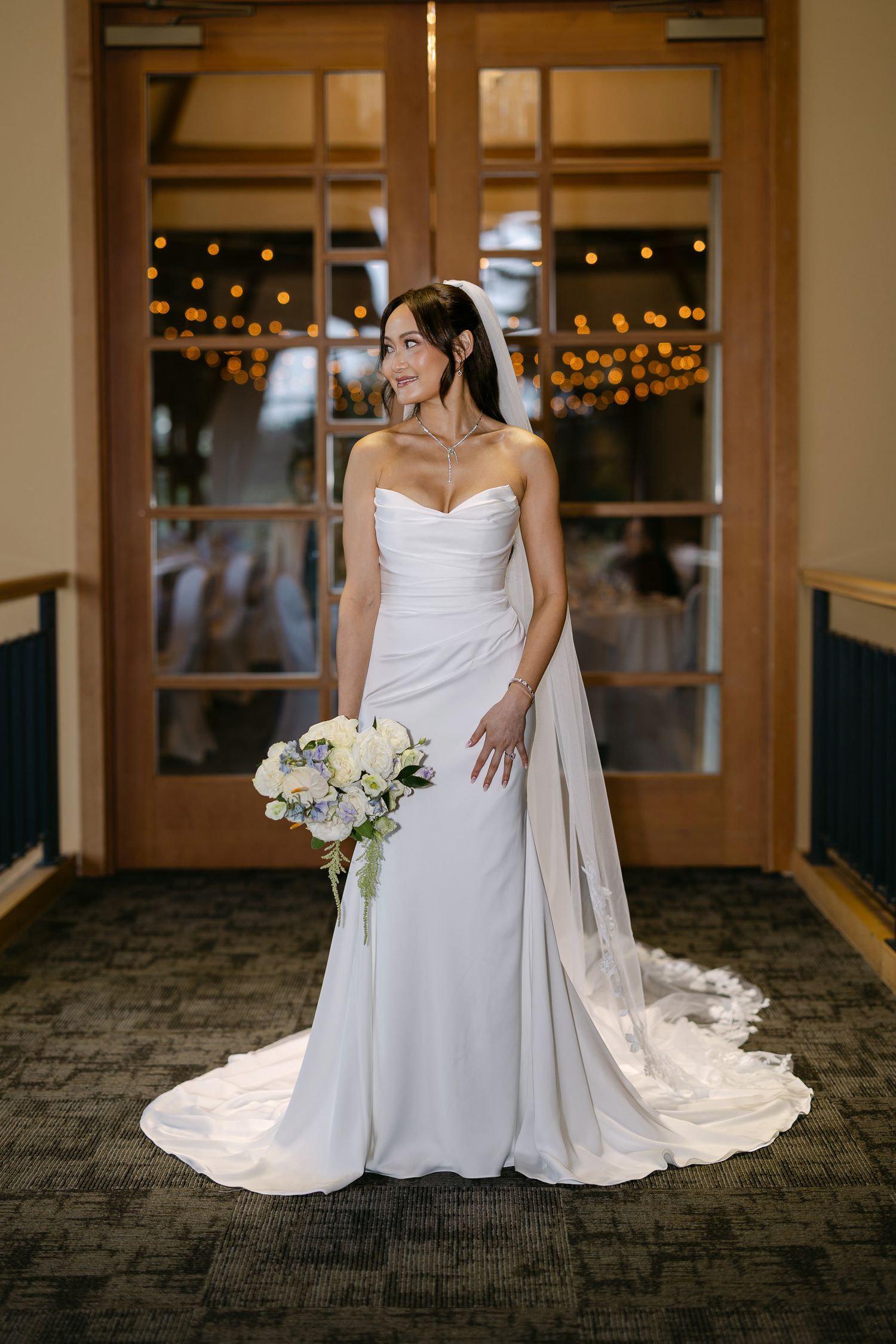 Bride in strapless white gown with lace-trimmed train poses holding white and blue bouquet.