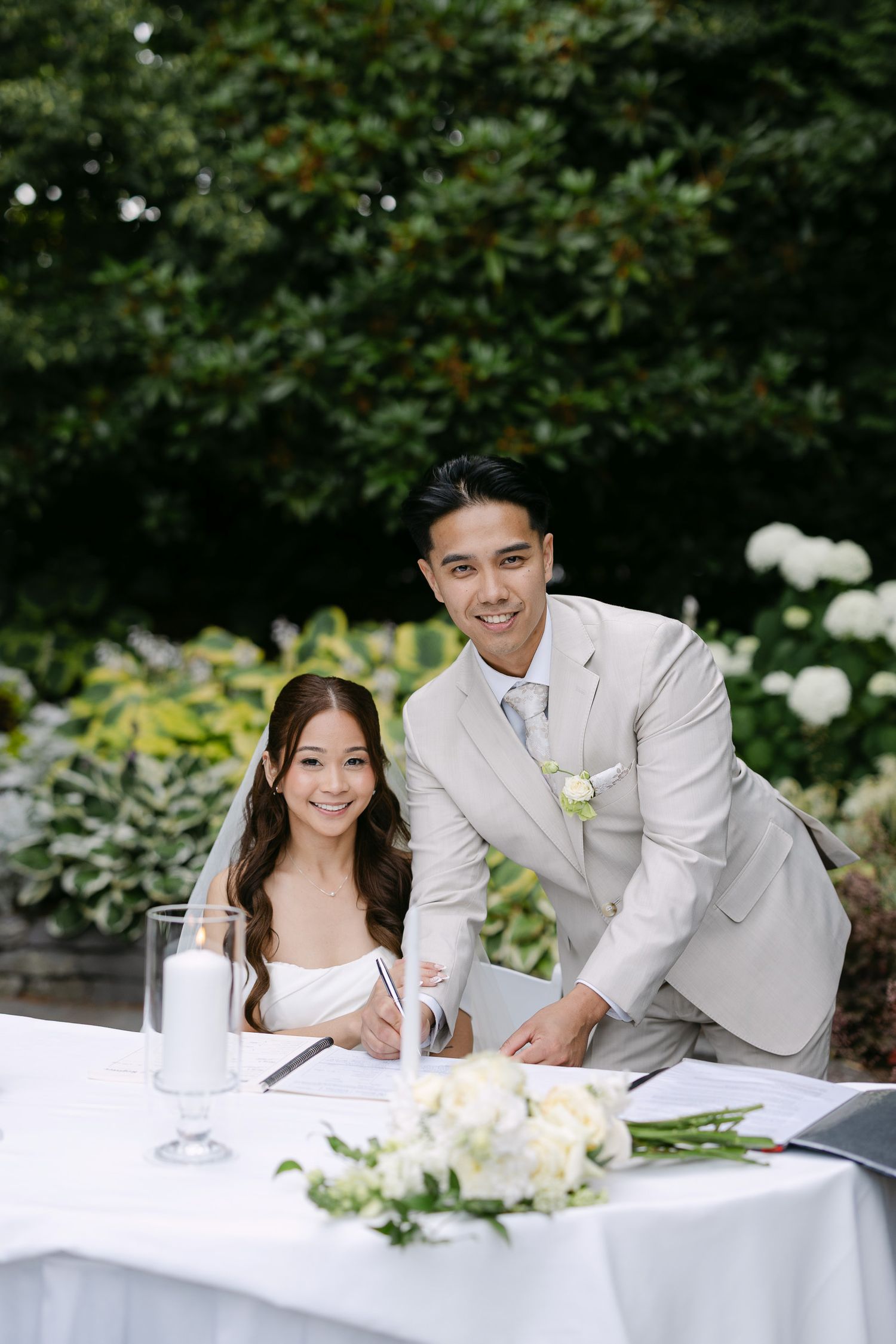 A bride and groom smile while signing their wedding registry at an outdoor ceremony table with white flowers.