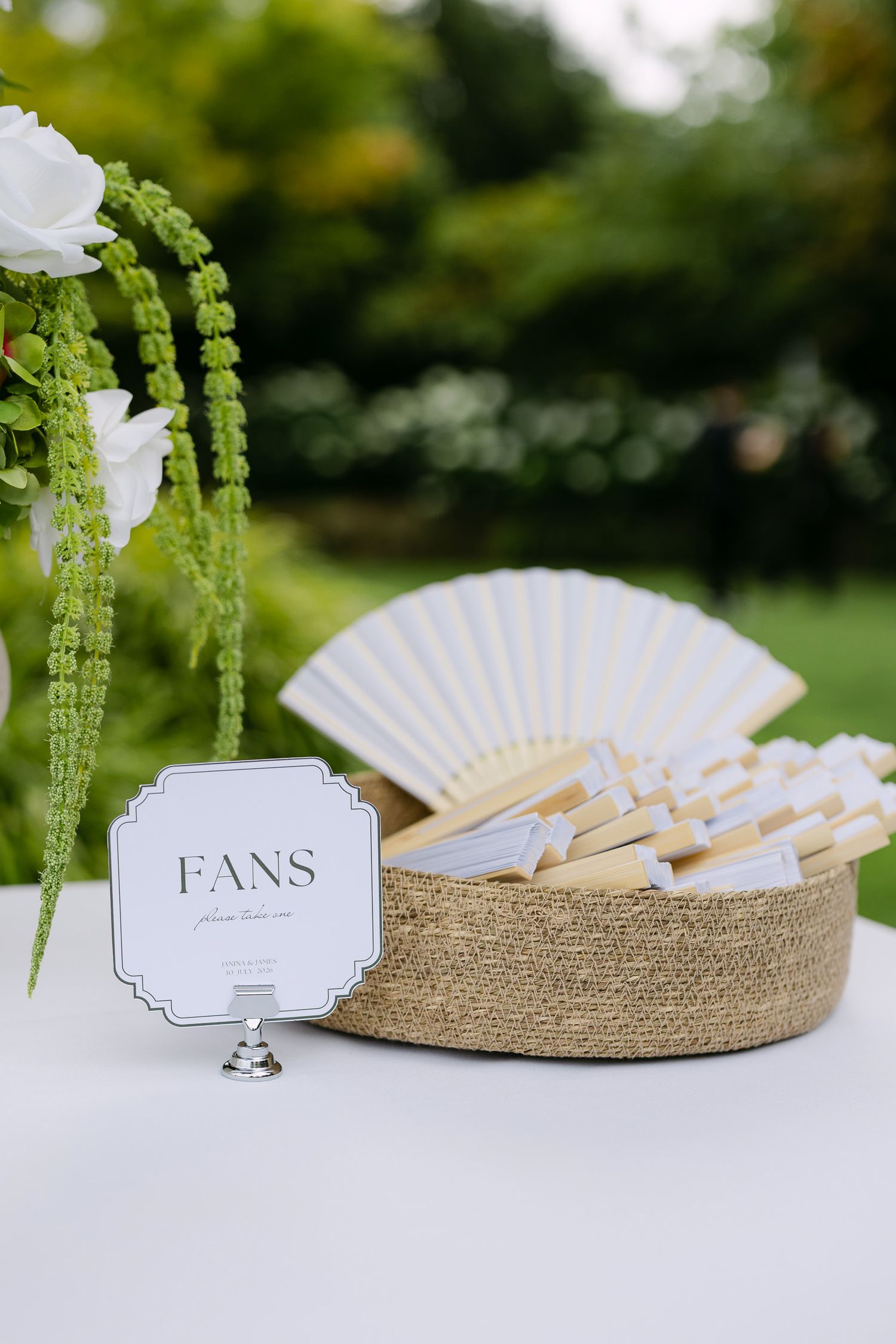 Wicker basket filled with white hand fans displayed on a table with a decorative sign at an outdoor wedding.