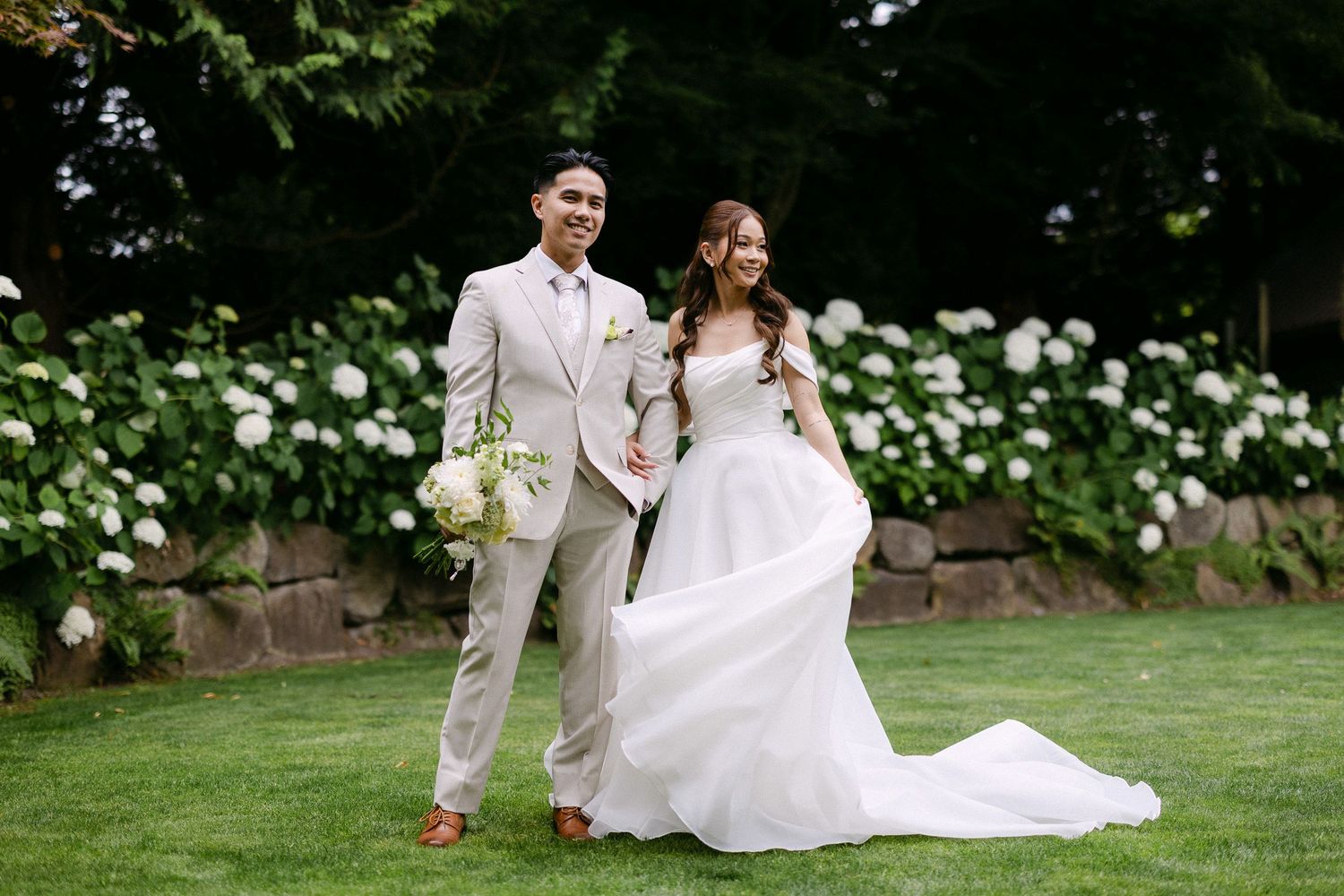Bride in white gown and groom in tan suit pose together on green lawn with white hydrangeas in background.