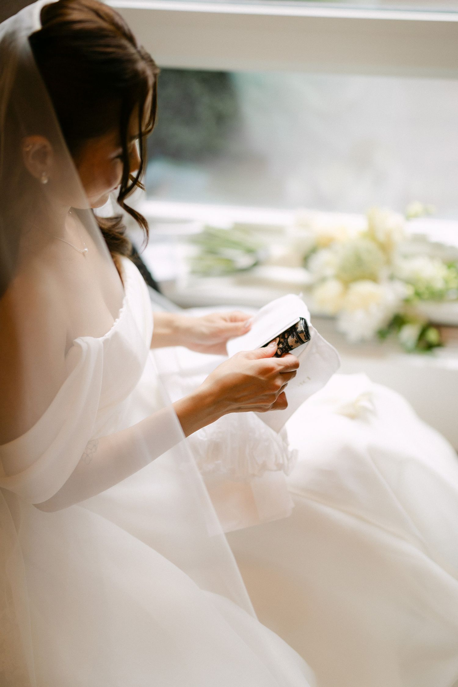 A bride in a white gown and veil reads a heartfelt letter by a window with a white floral bouquet nearby.