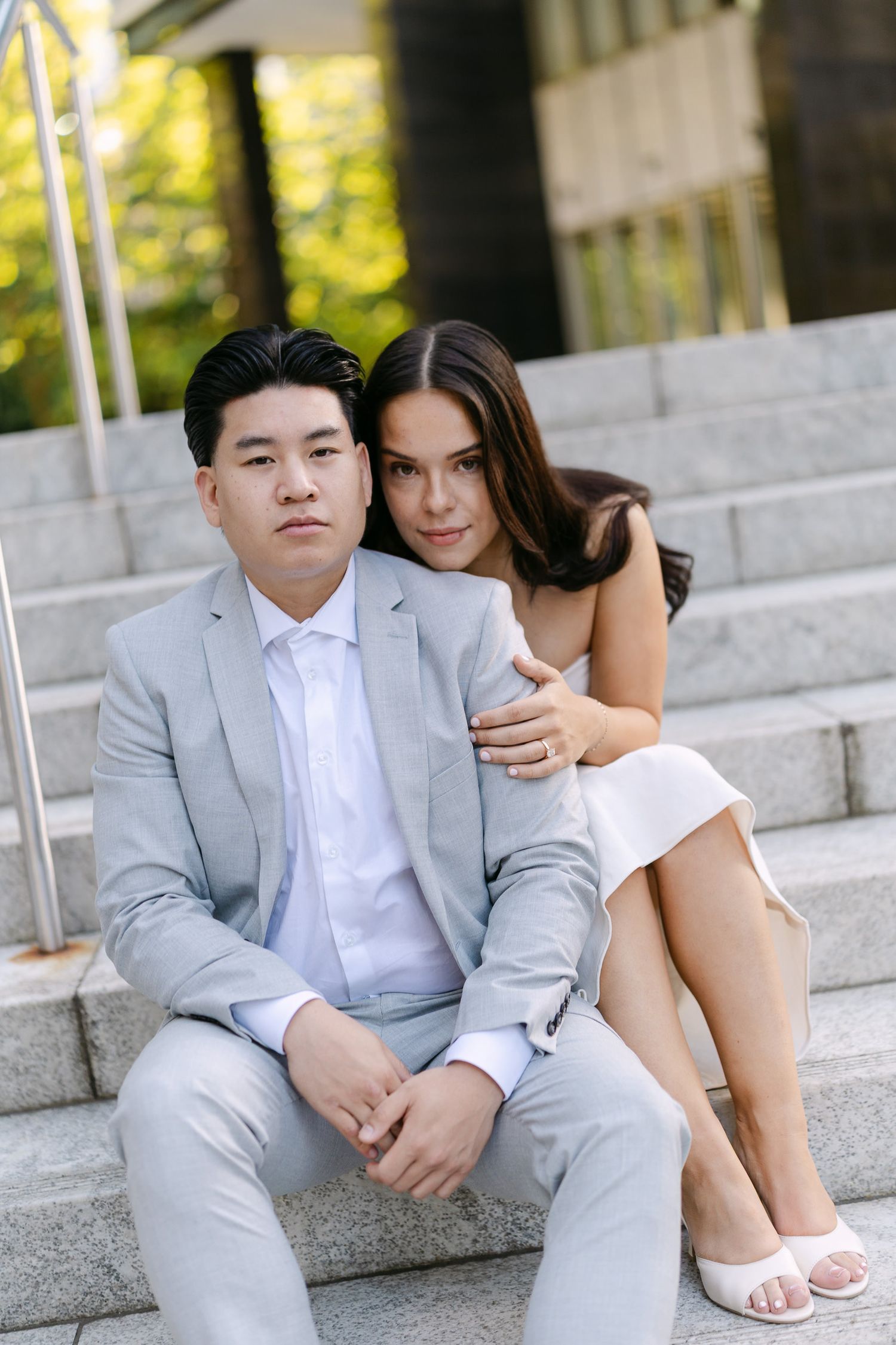 Couple in a light gray suit and white dress sit close together on outdoor stone steps during an engagement photo session.