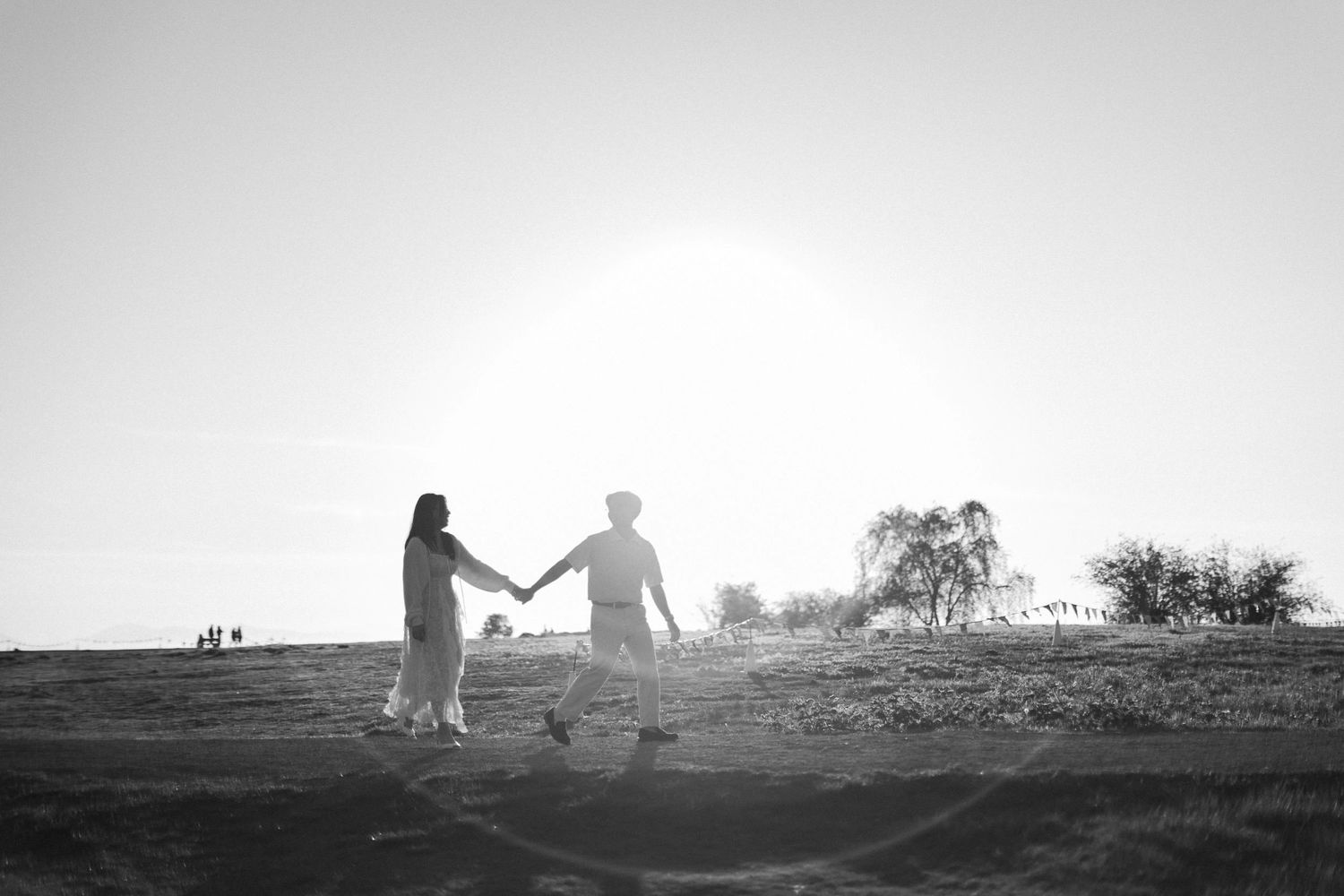 Black and white silhouette of a couple holding hands and walking across an open field toward the setting sun.