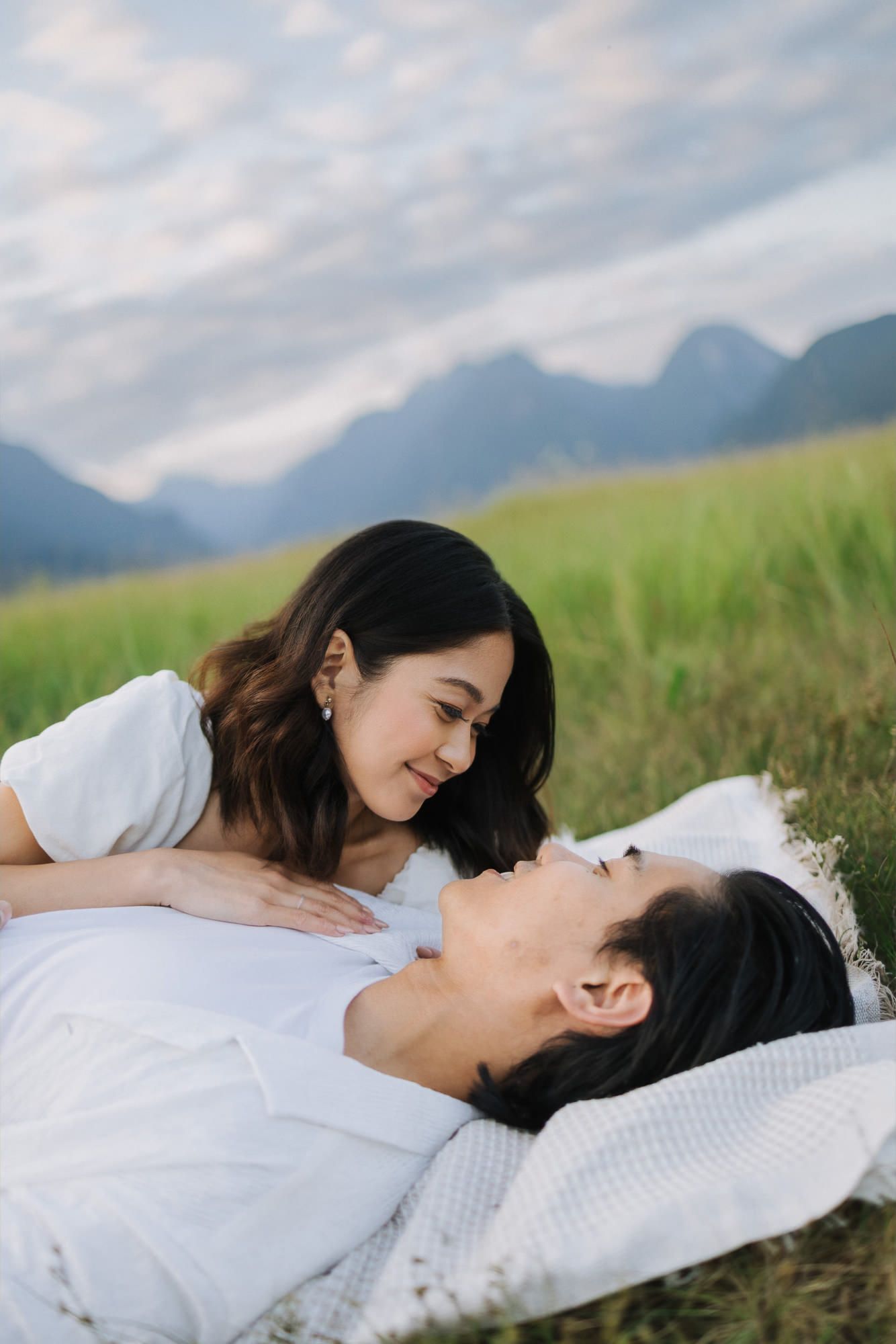 Smiling woman in white leans over her partner as they relax on a blanket in a green meadow with mountains behind.
