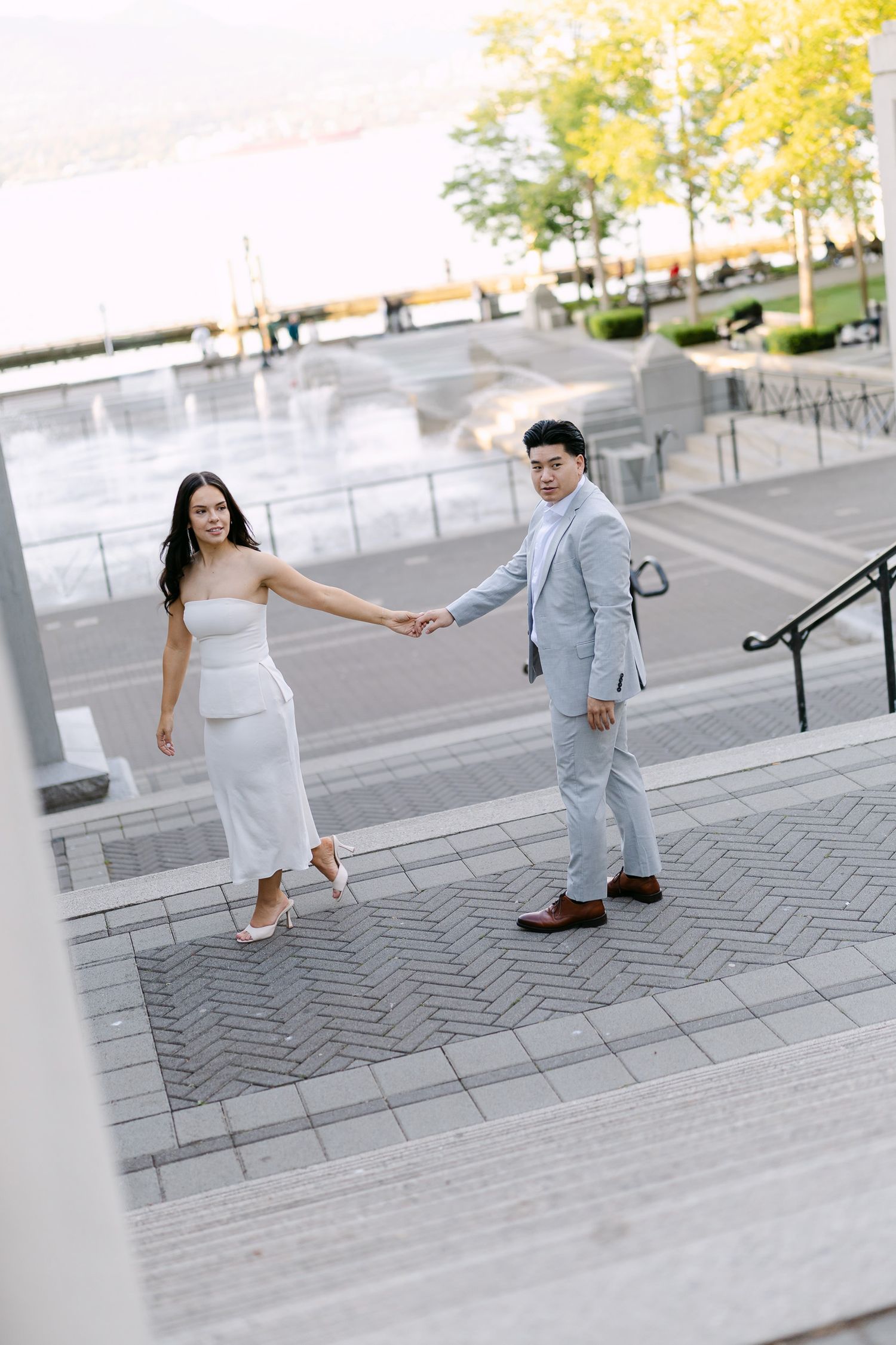 Couple holding hands walking down waterfront stairs, she in a white dress and he in a light gray suit, during an engagement photo session.