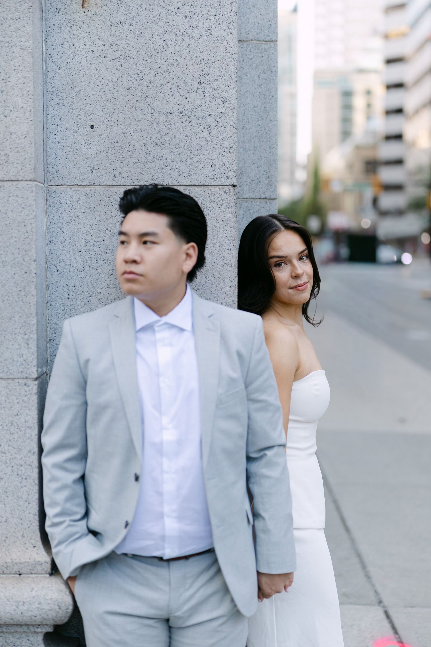 Couple in engagement photos beside a granite pillar on a city street, a man in a gray suit and a woman in a white strapless dress.