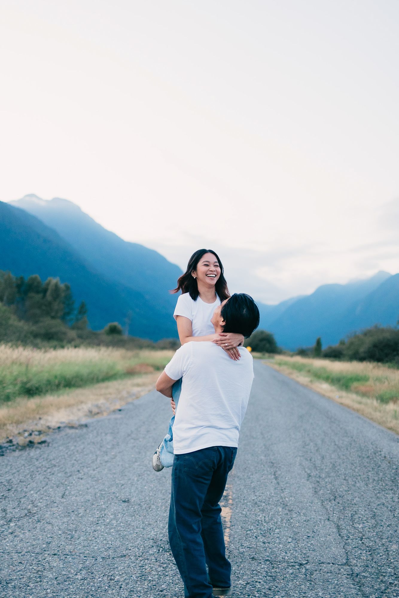 Smiling woman in a white top is lifted by her partner on a quiet country road with mountains in the background.