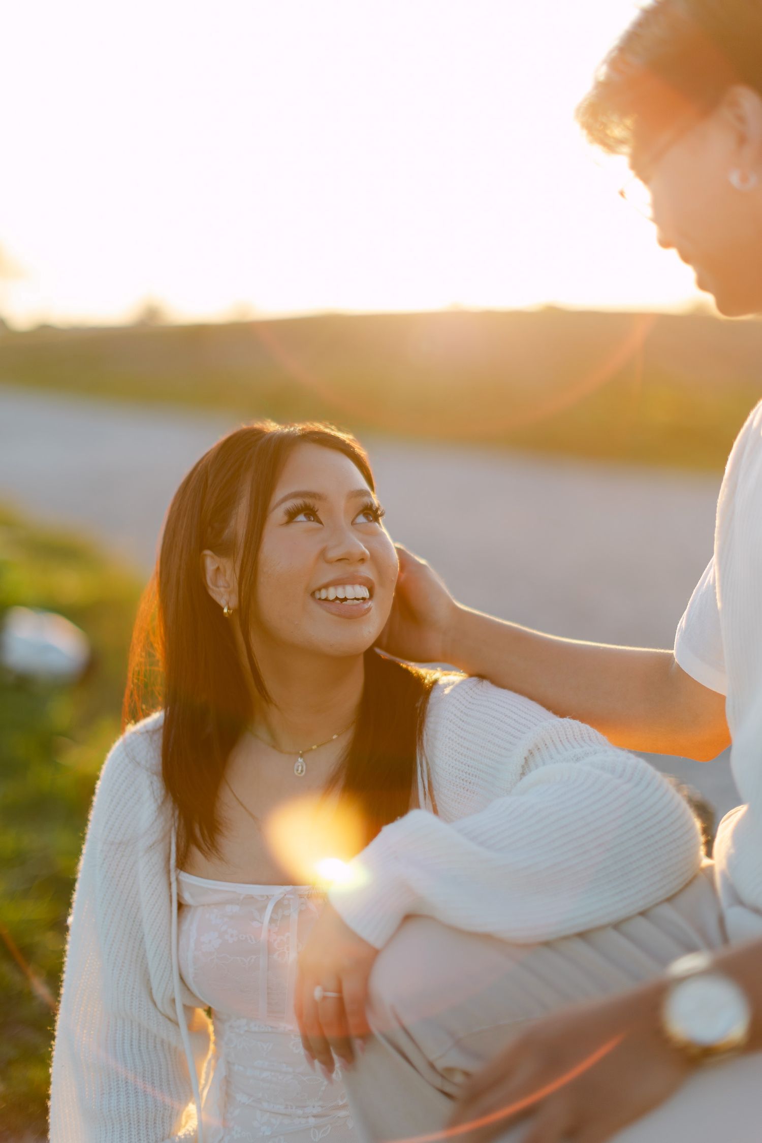 Smiling woman in a white cardigan gazes up lovingly at her partner during a golden hour sunset engagement photo session outdoors.