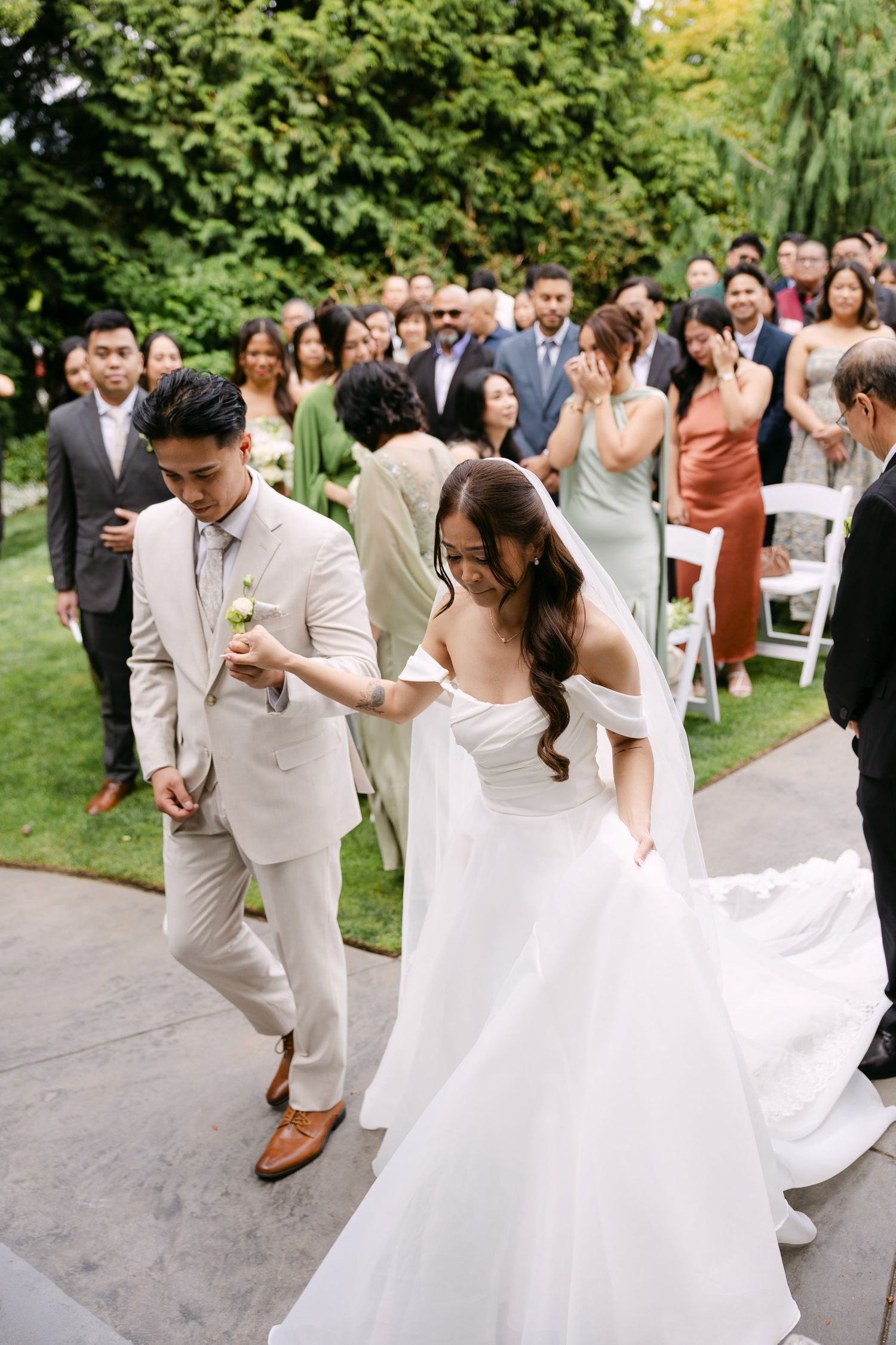 Newlywed couple walking down aisle after outdoor wedding ceremony with guests celebrating behind them.