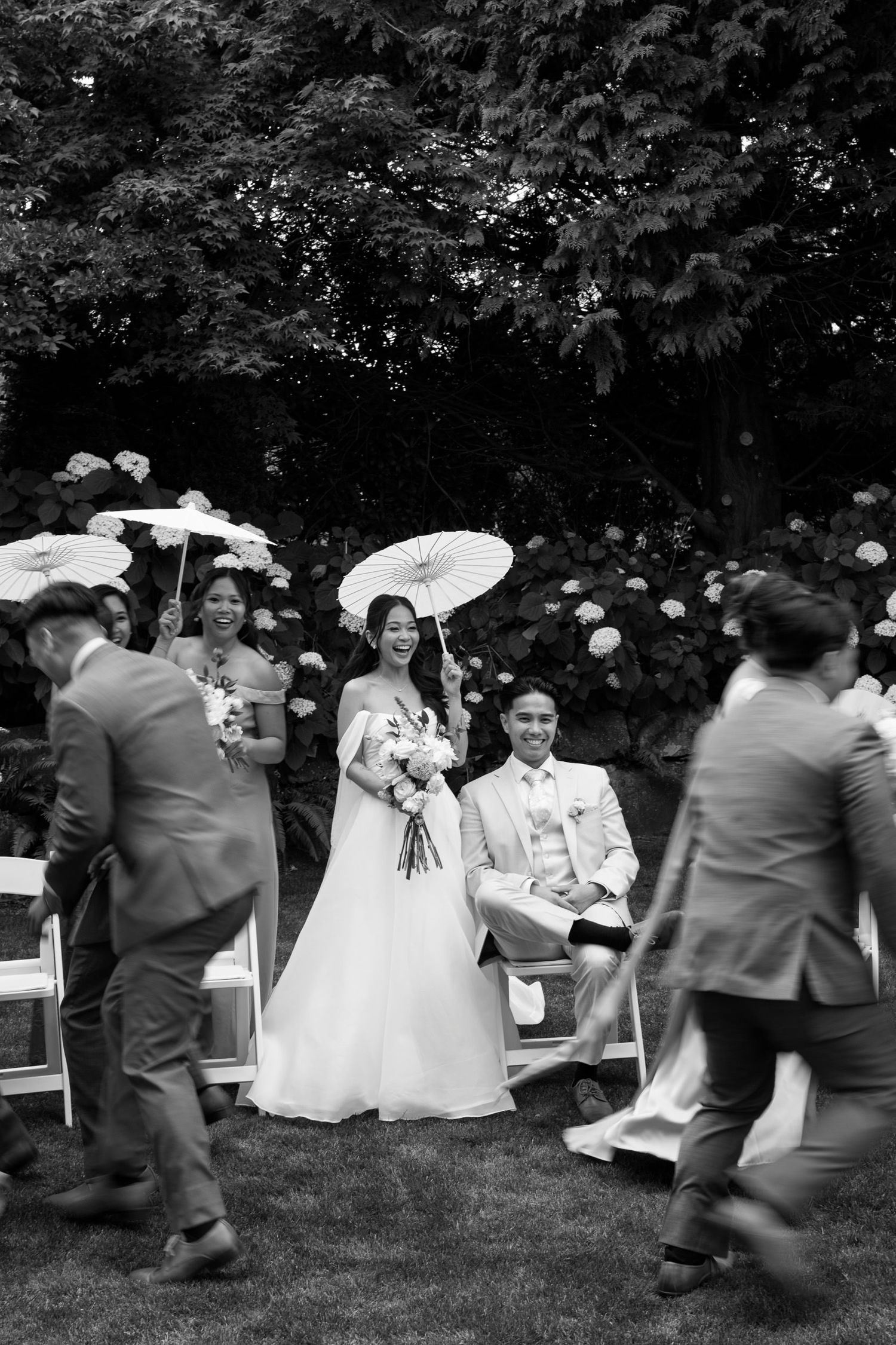 Black and white outdoor wedding ceremony with bride holding parasol, groom seated, and guests walking past floral backdrop.