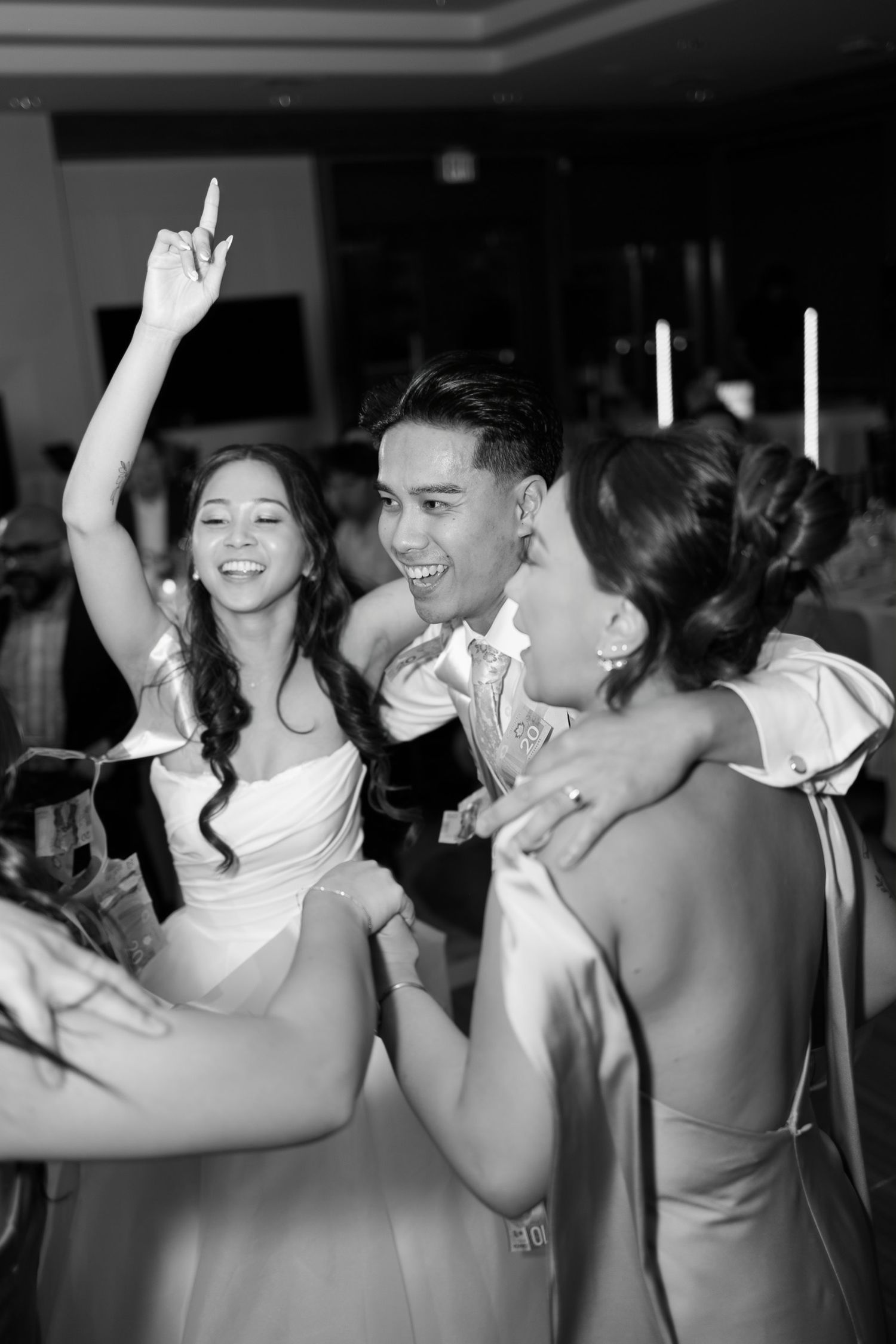 Black and white photo of a bride, groom, and bridesmaid joyfully dancing and celebrating at a wedding reception.