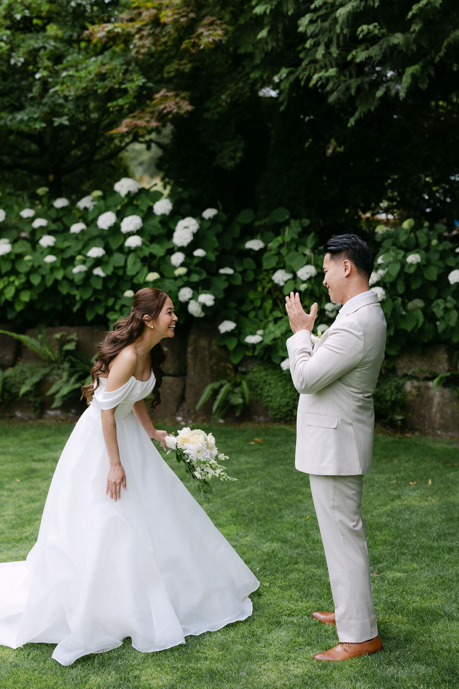 Bride in white off-shoulder gown holds bouquet as groom in tan suit reacts emotionally during first look in garden.