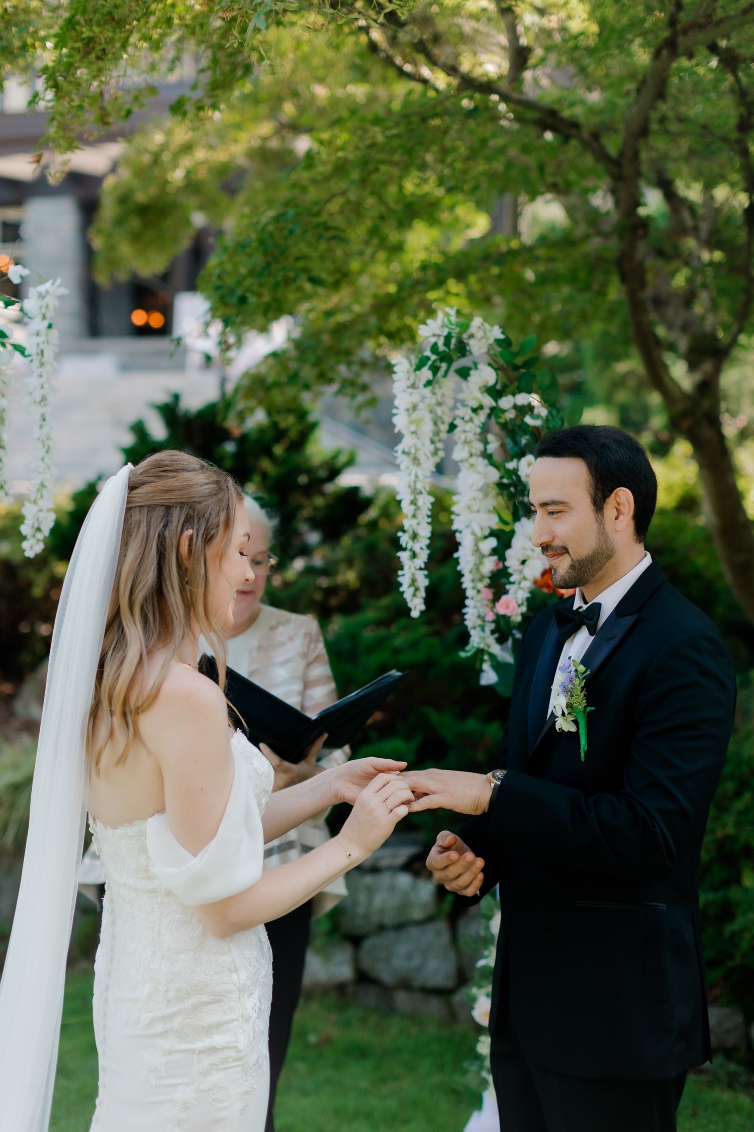 A couple exchanges rings during an outdoor wedding ceremony with white floral decorations and lush greenery in the background.