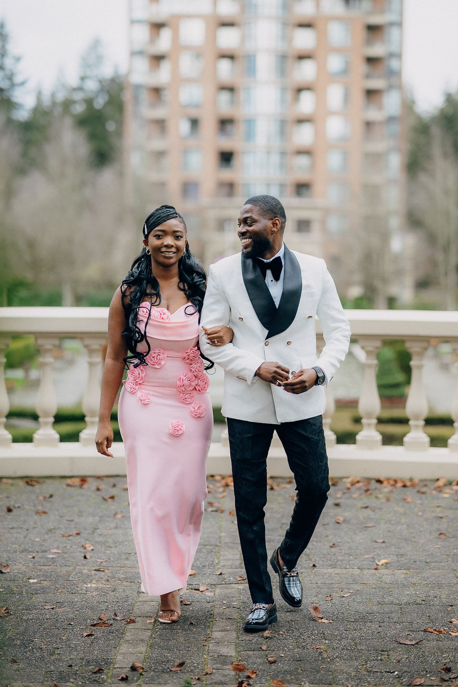 A couple in formal attire, with pink dress and white tuxedo jacket, dance playfully together outdoors.