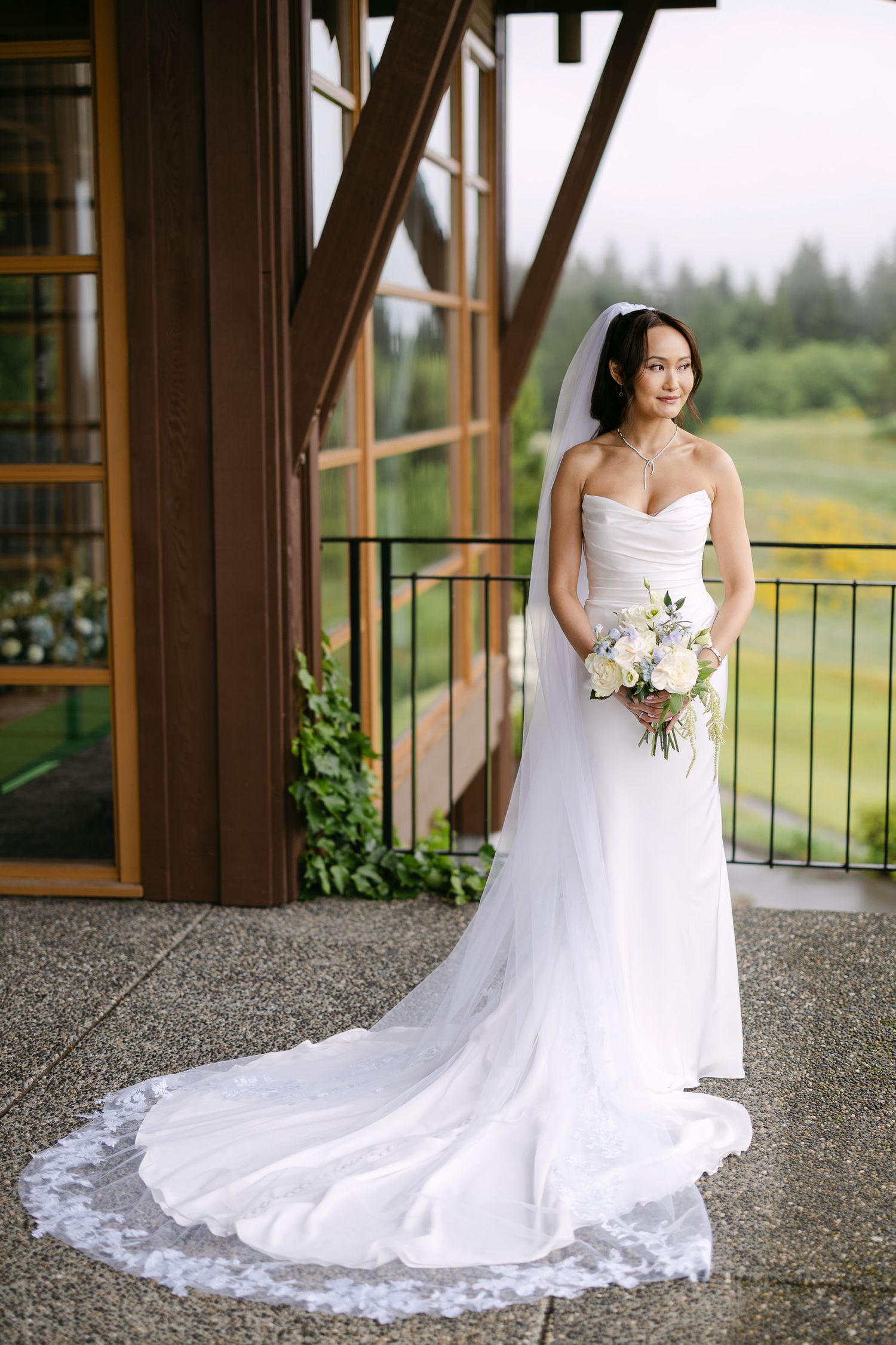 Bride in strapless white gown with cathedral veil holds bouquet on outdoor balcony.