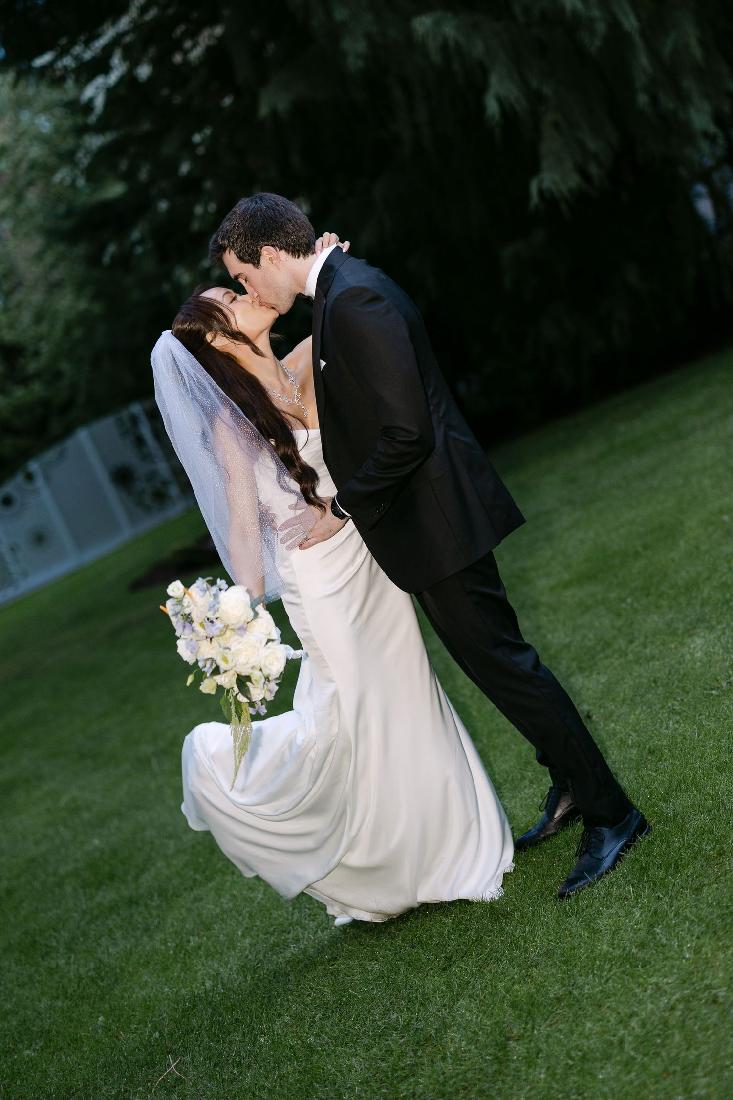 Bride and groom sharing romantic dip kiss on green lawn, bride in white gown holding bouquet.