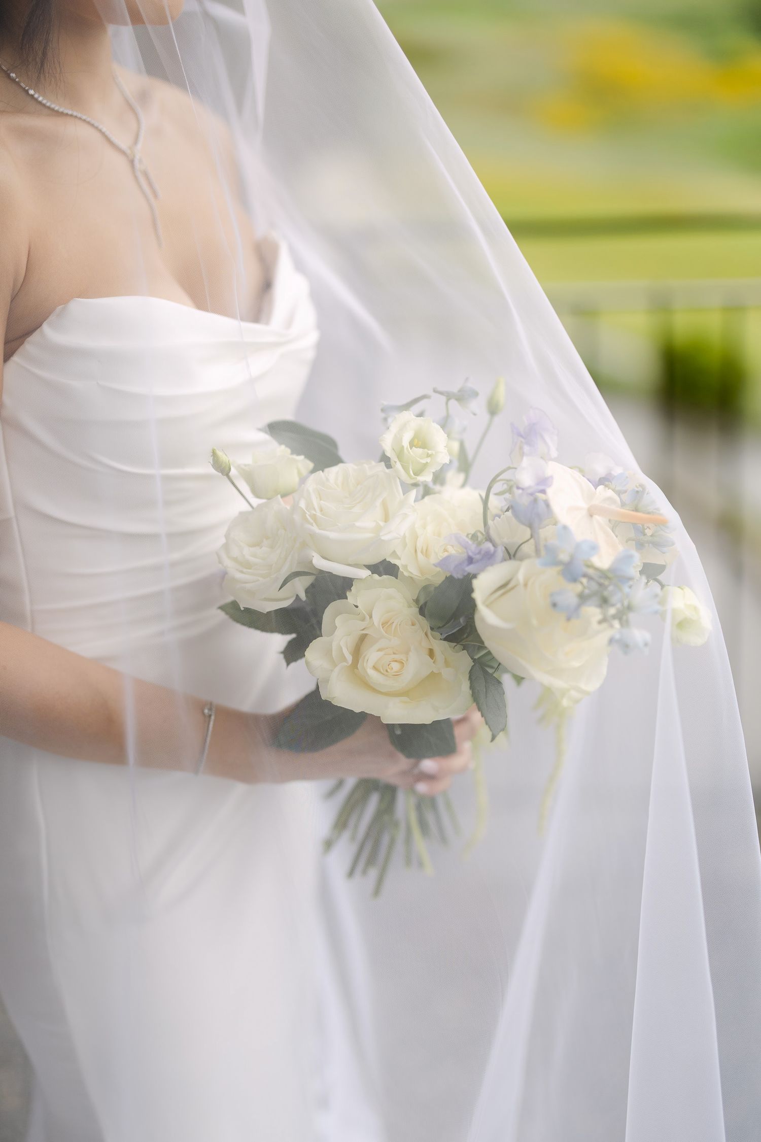 Bride in white strapless gown holding elegant bouquet of white roses and blue flowers, draped in flowing veil.