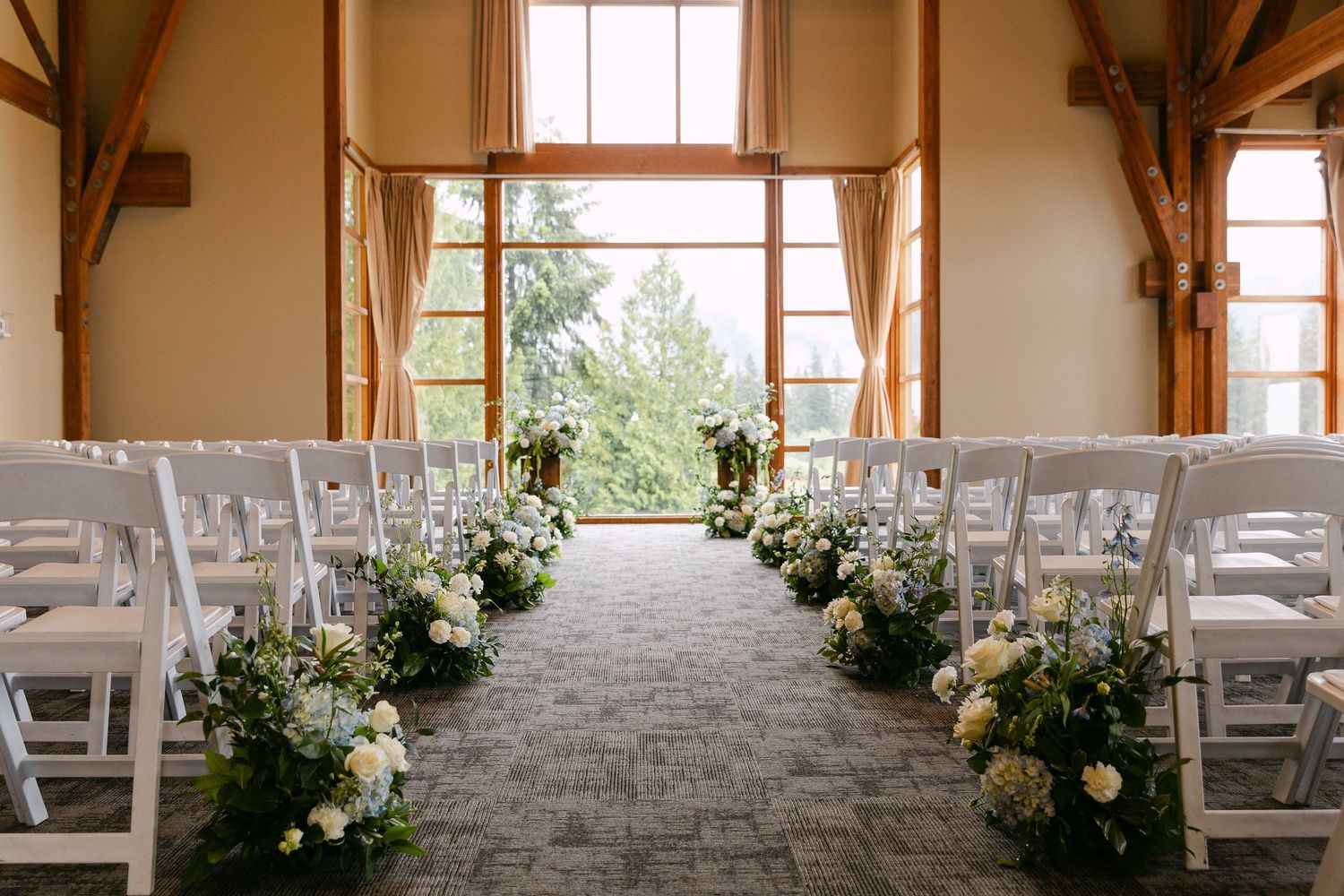 Elegant indoor wedding ceremony aisle with white chairs and lush floral arrangements leading to large windows.