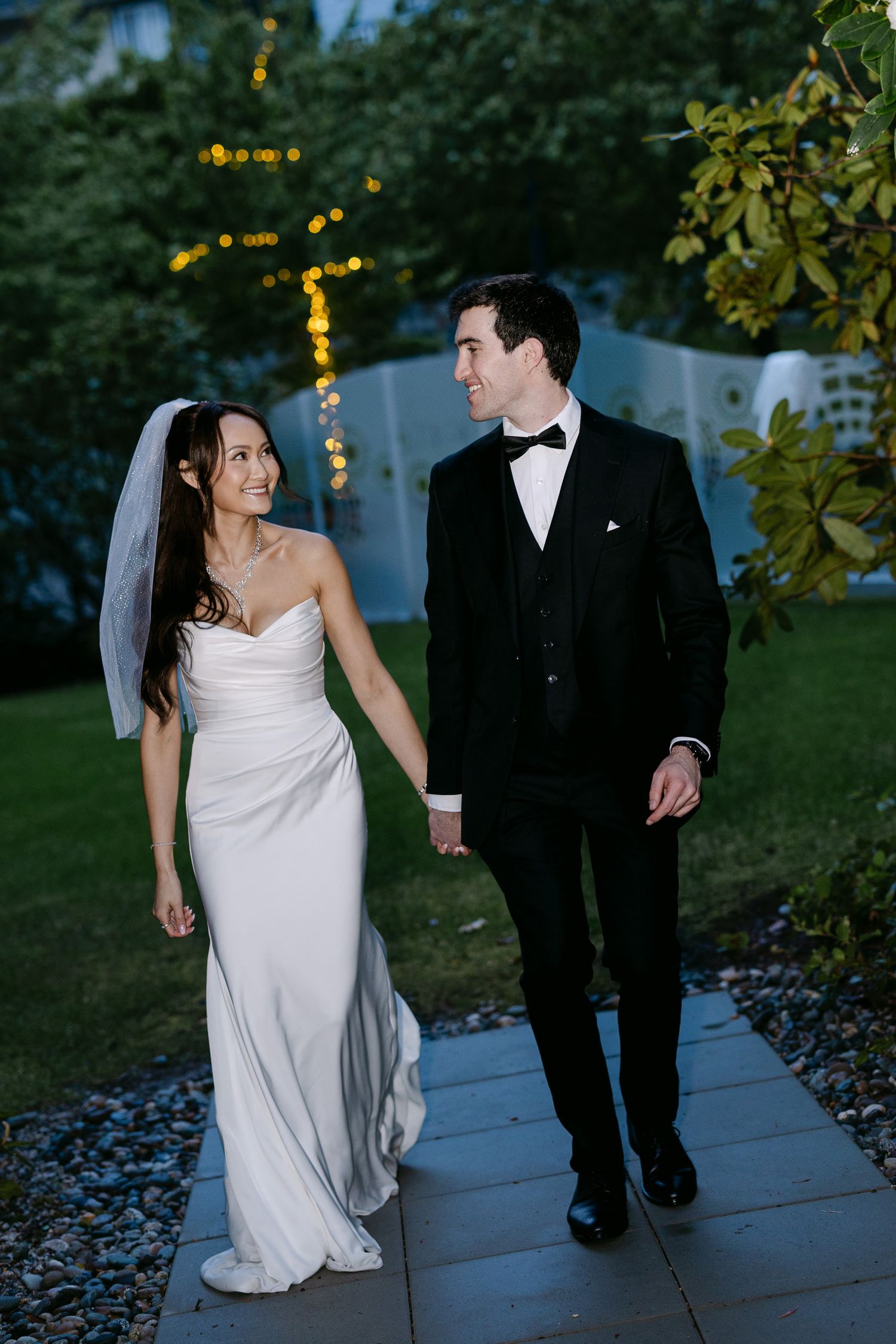 Bride and groom walk hand in hand along garden path at dusk with string lights.