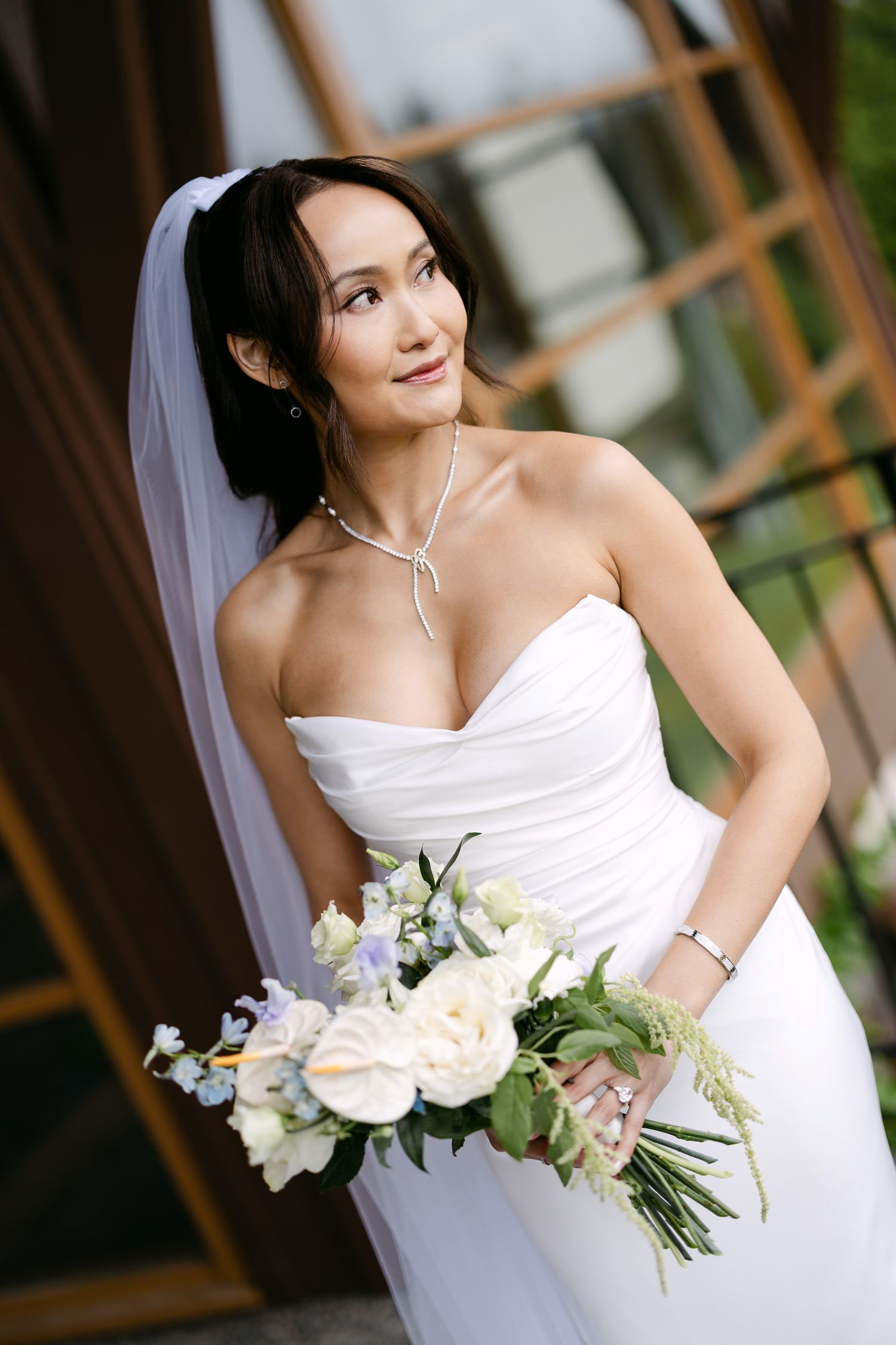 Elegant bride in strapless white gown holding bouquet of white and blue flowers, wearing veil and pearl necklace.