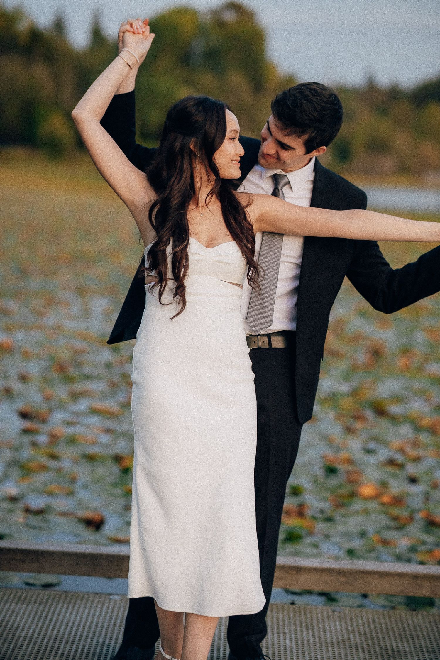 A couple in formal attire dancing together on a dock surrounded by autumn leaves on the water.