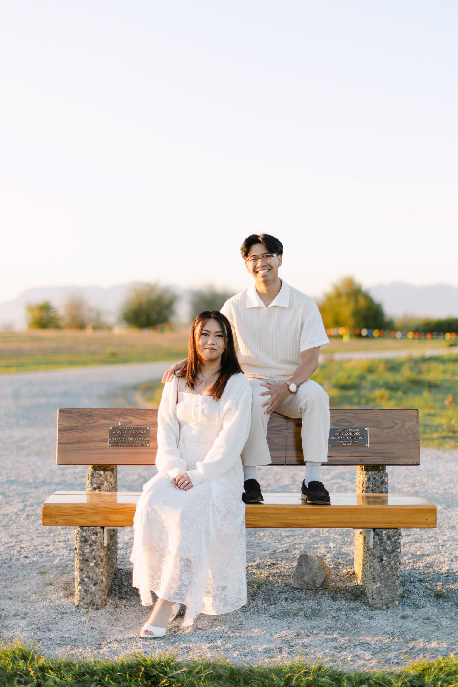 Smiling couple posing on a wooden park bench at sunset, she in a white lace dress seated in front, he in a cream polo behind.