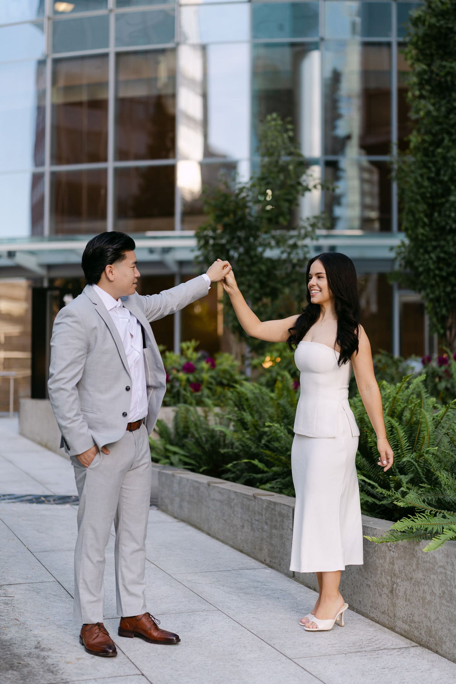 Couple in elegant attire share a romantic dance on a modern city plaza, with the man in a gray suit twirling the woman in a white dress.