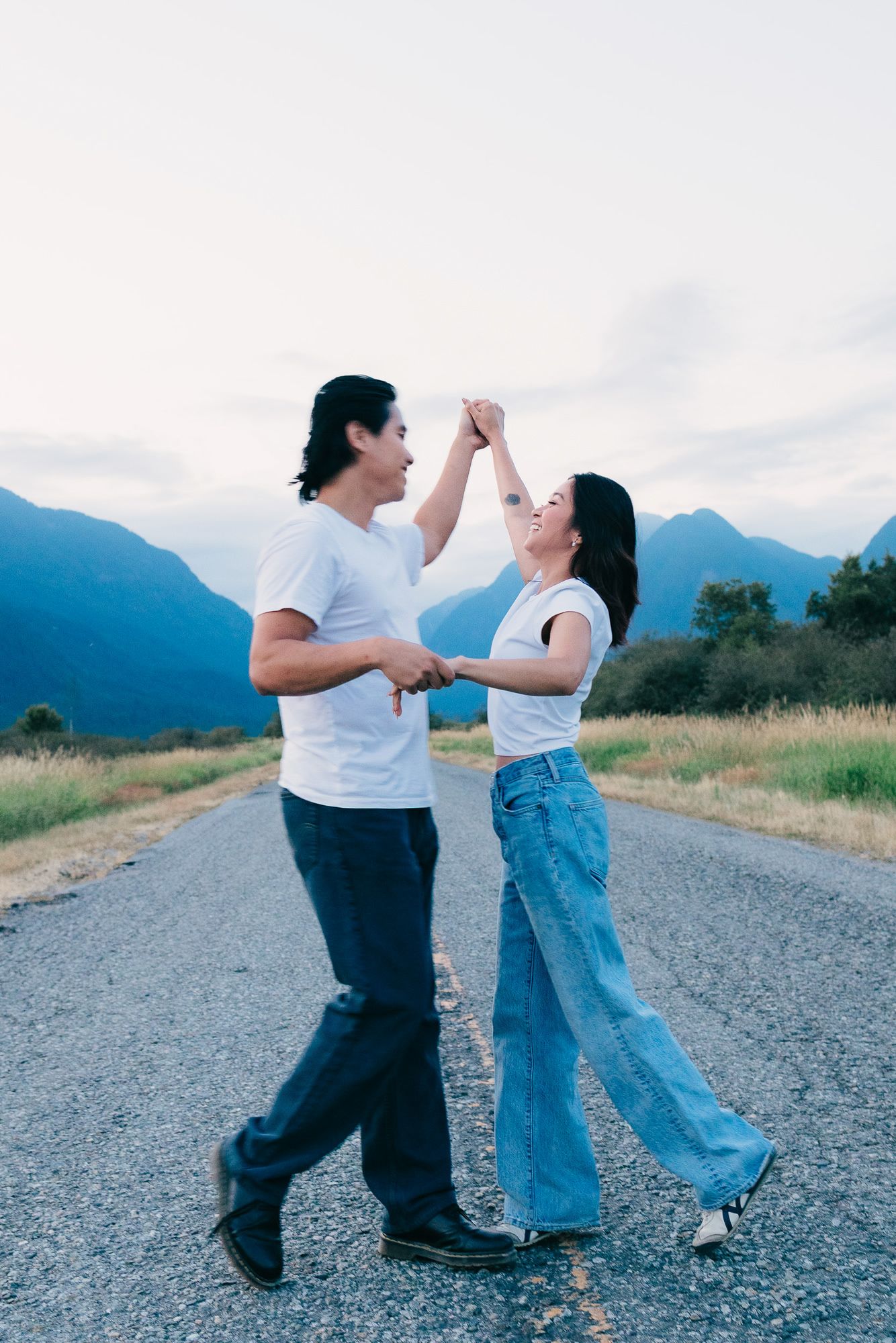 Smiling couple dancing hand in hand on a quiet paved road with mountains in the background at dusk.