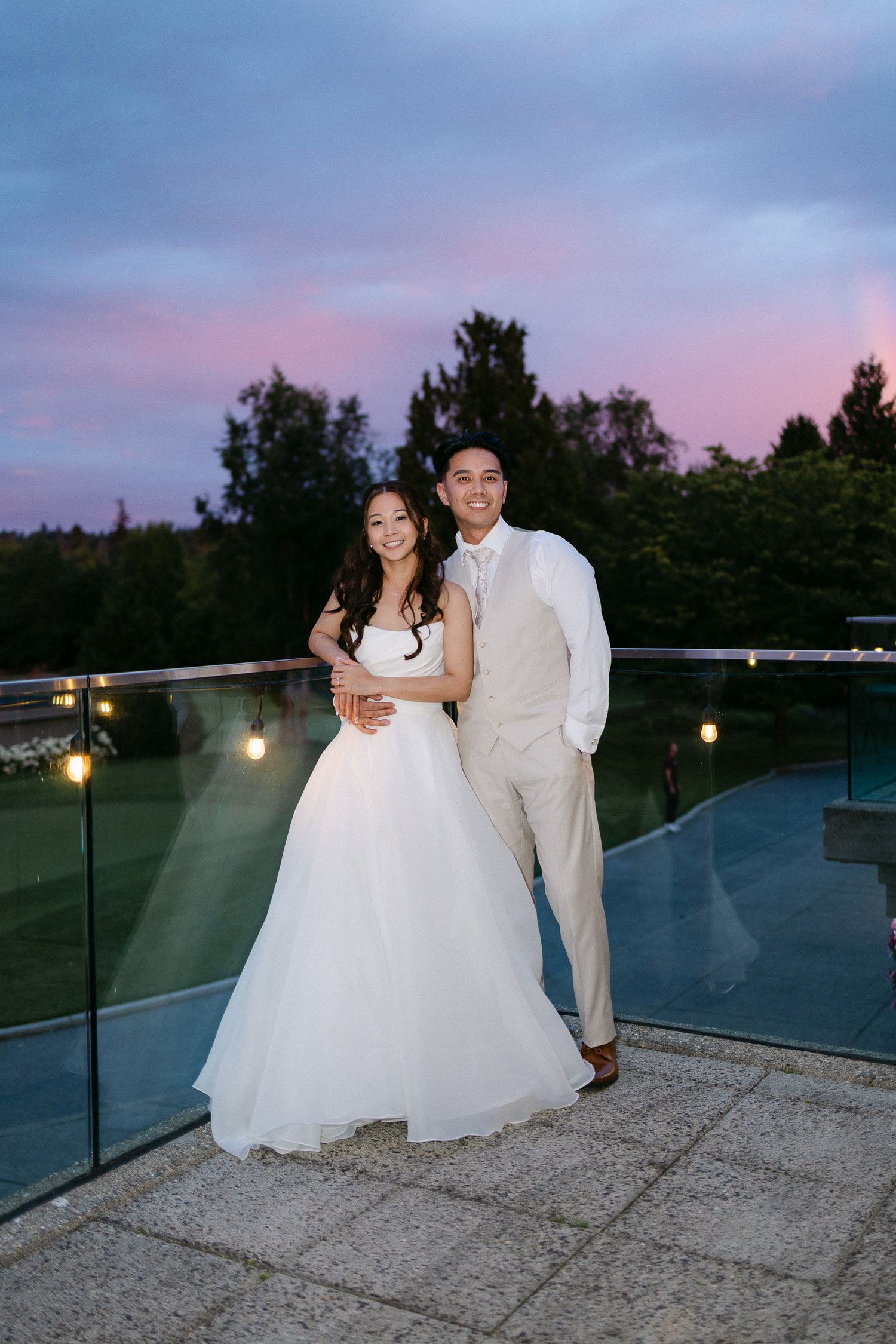 Bride in white gown and groom in tan suit embrace on terrace at sunset with pink and purple sky.