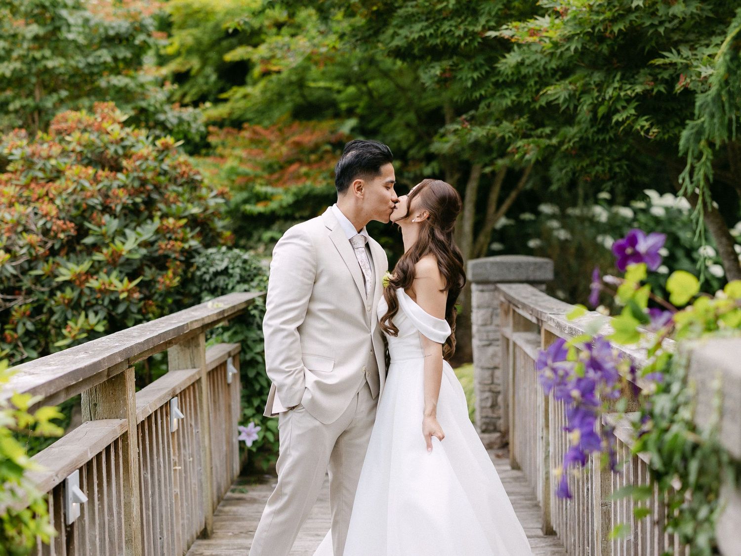 A couple shares a romantic kiss on a wooden bridge surrounded by lush greenery in a garden wedding setting.