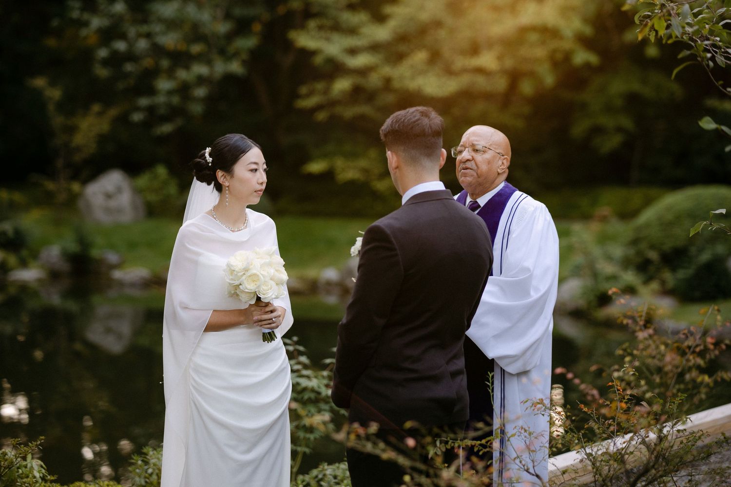 Bride and groom exchange vows with officiant in a serene garden wedding ceremony near a tranquil pond.