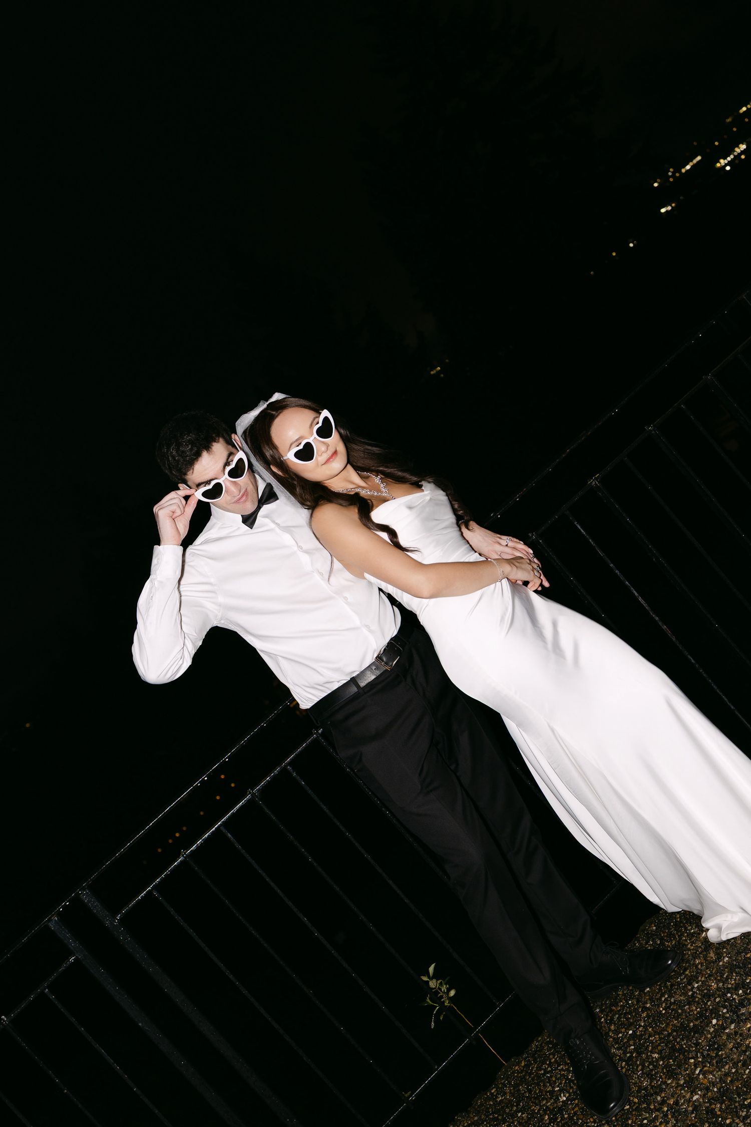 Bride and groom in white outfits and heart-shaped sunglasses pose playfully at night near a railing.